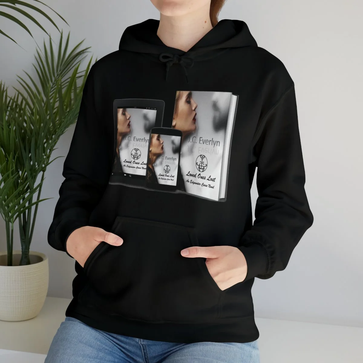 Person wearing a black hoodie with a graphic print of a woman’s profile and books, standing next to a potted plant.