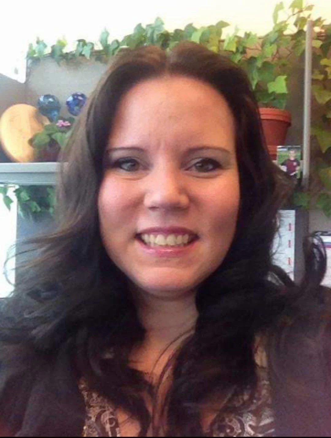 A smiling woman with long dark hair, sitting in an indoor space with plants and decorative items in the background.
