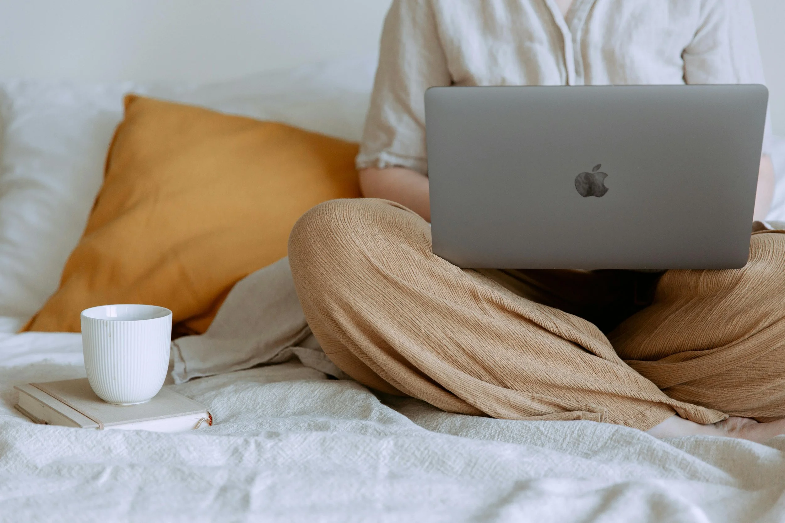 Person sitting cross-legged on a bed with a laptop, book, and mug nearby