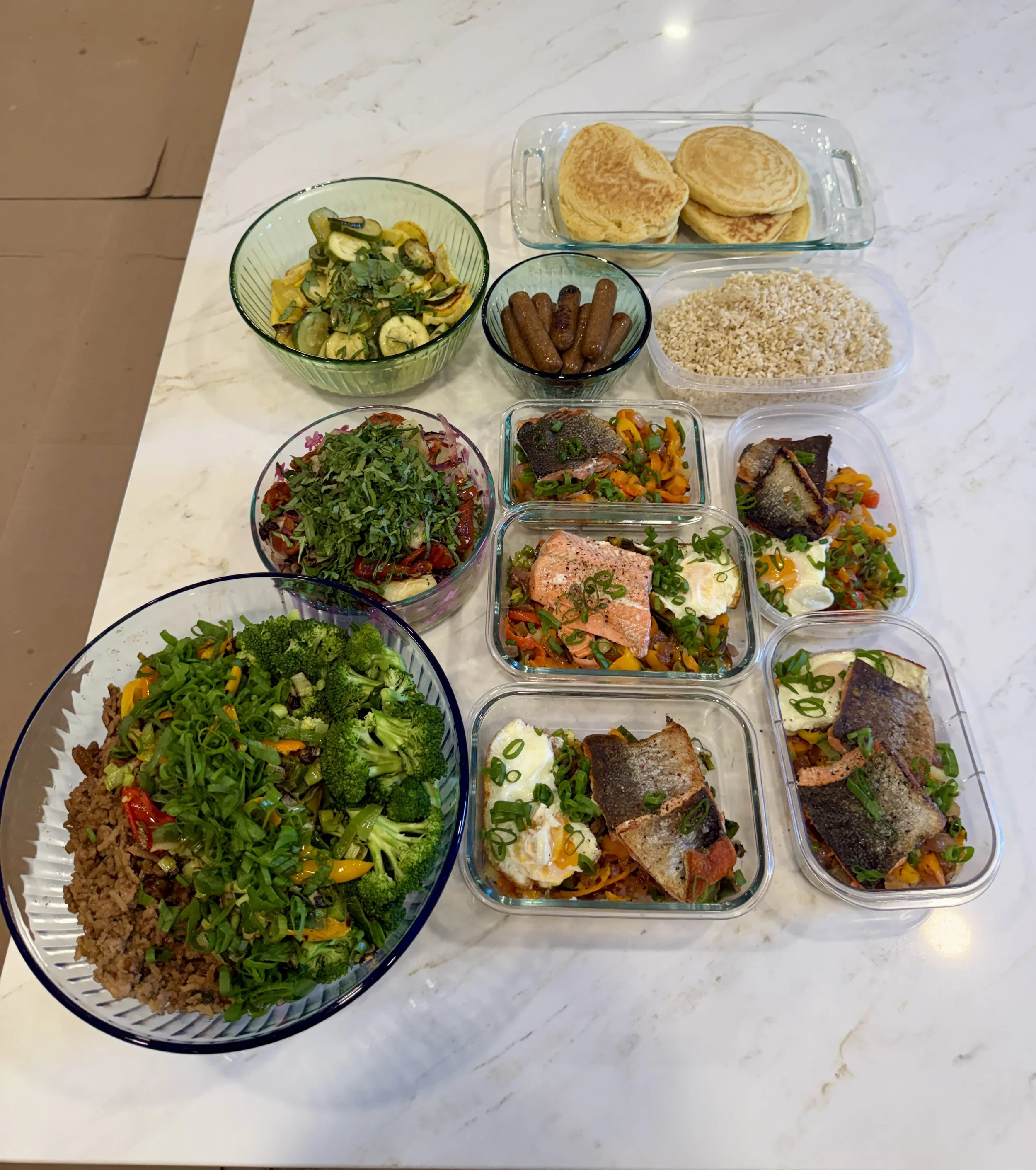 Assorted dishes on a marble countertop, including a large bowl of salad with greens and vegetables, a container of cooked ground meat with chopped green onions, and several small containers with cooked fish, grilled meats, and seasoned rice.