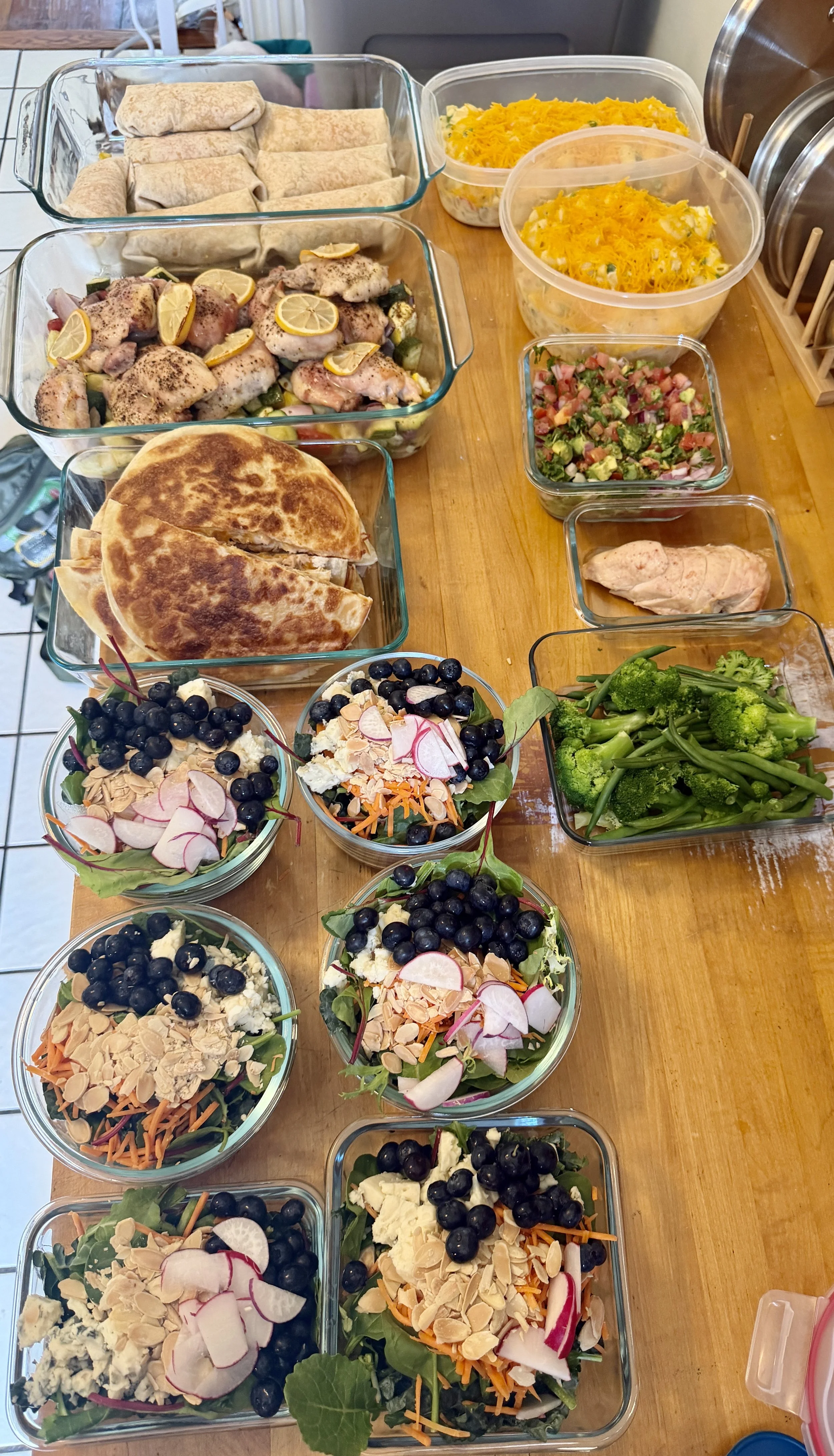 Prepared salads, grilled chicken with lemon slices, shredded cheese, rolled tortillas, and broccoli on a wooden table, ready for a meal or buffet.