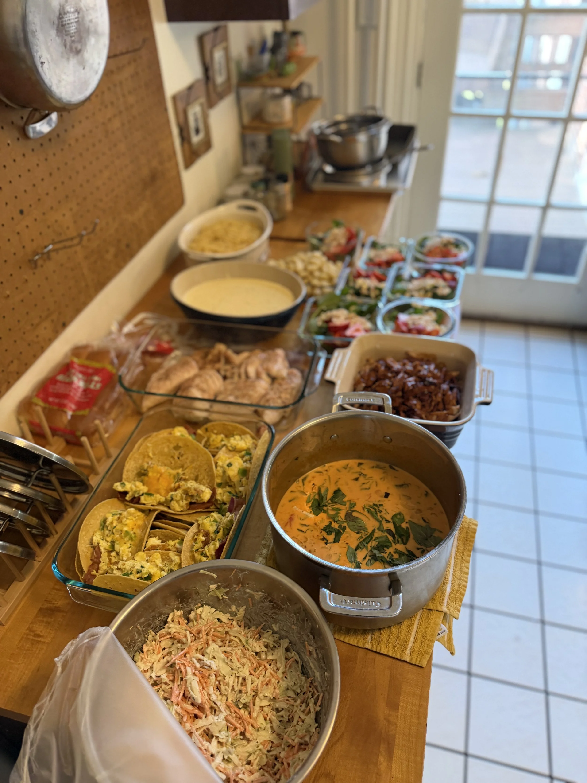 Assorted dishes on a kitchen counter, including potato salad, shrimp salad, taco fillings, and a large pot of soup with herbs. There are bowls, utensils, and a remote control nearby.