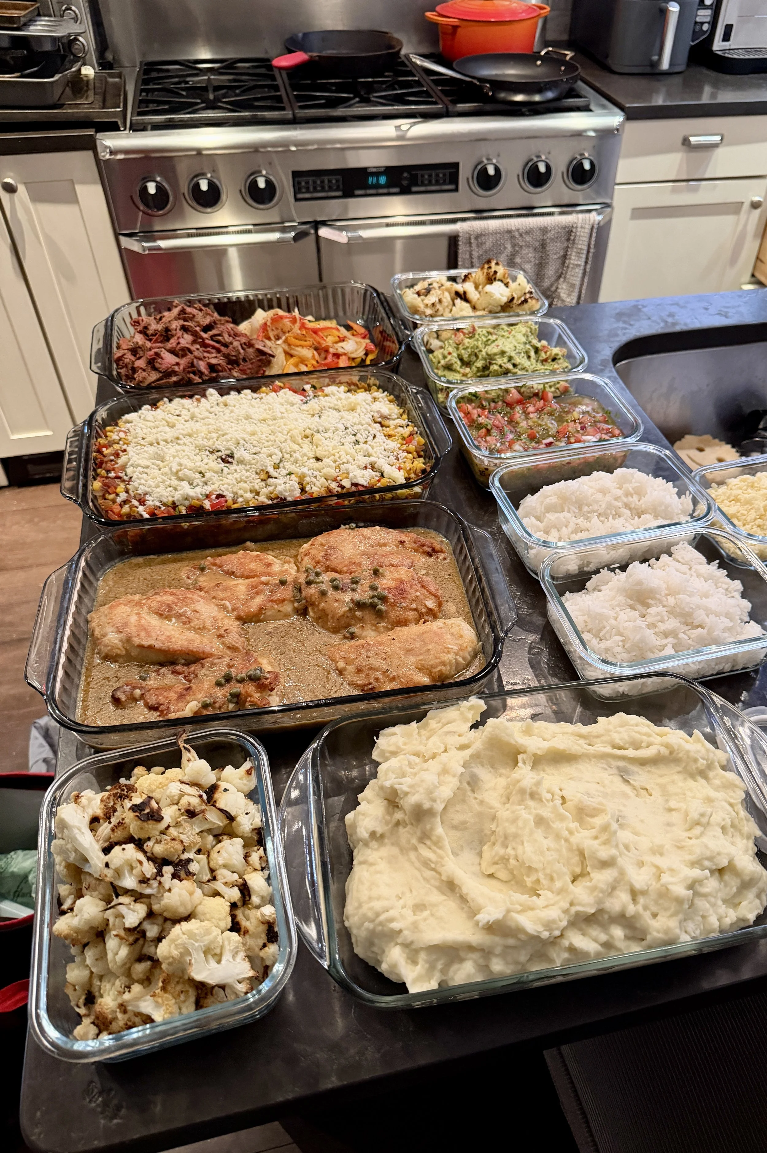 A kitchen countertop with multiple glass dishes filled with various foods, including mashed potatoes, a casserole topped with cheese, cooked beef, chopped vegetables, guacamole, salsa, and cauliflower salad. In the background, there is a stove, pans,