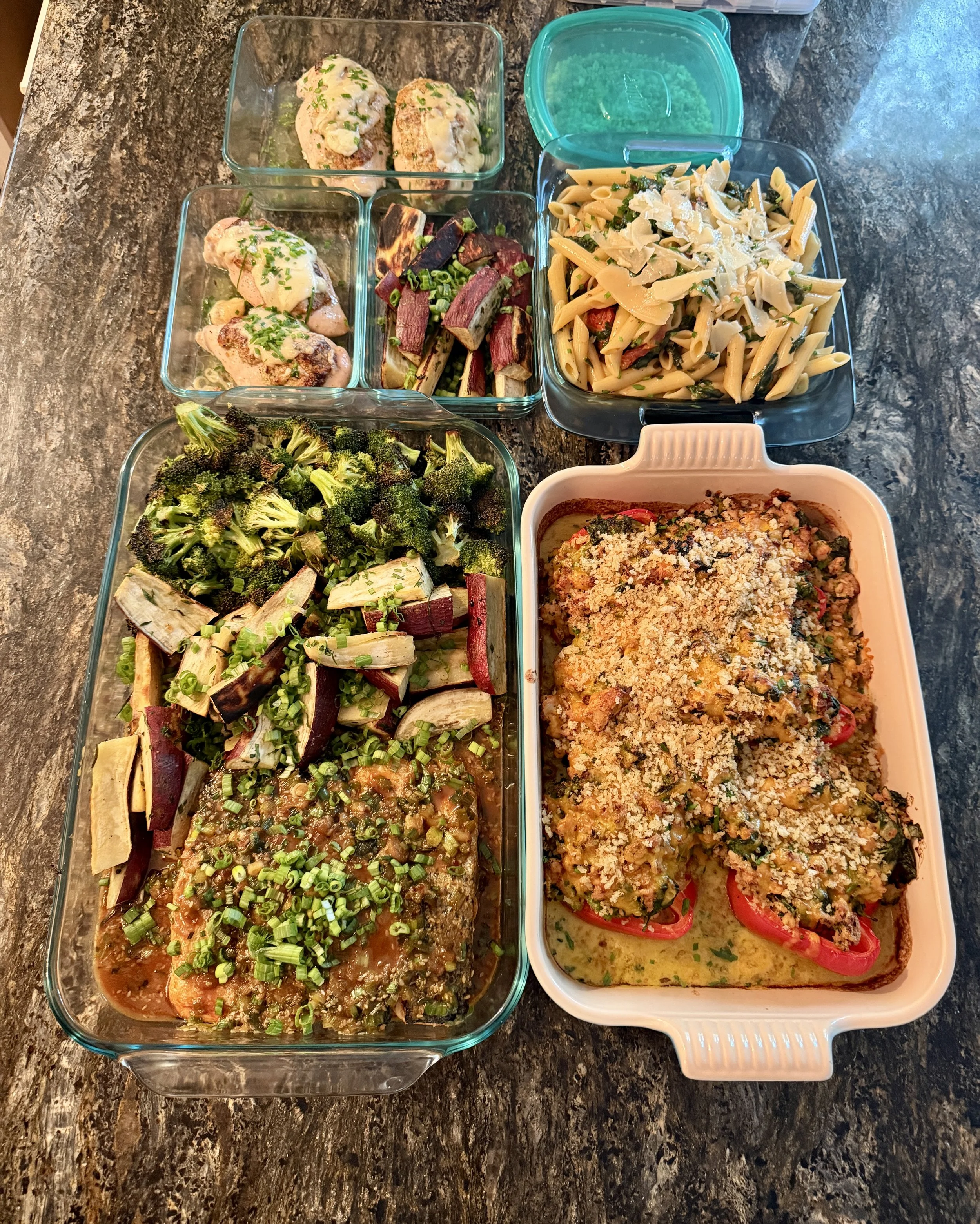 An assortment of baked dishes and vegetables on a dark marble countertop. Contains a casserole with stuffed bell peppers topped with breadcrumbs, roasted broccoli and eggplant, a tray of baked fish with tomato, a bowl of pasta with cheese, and smalle