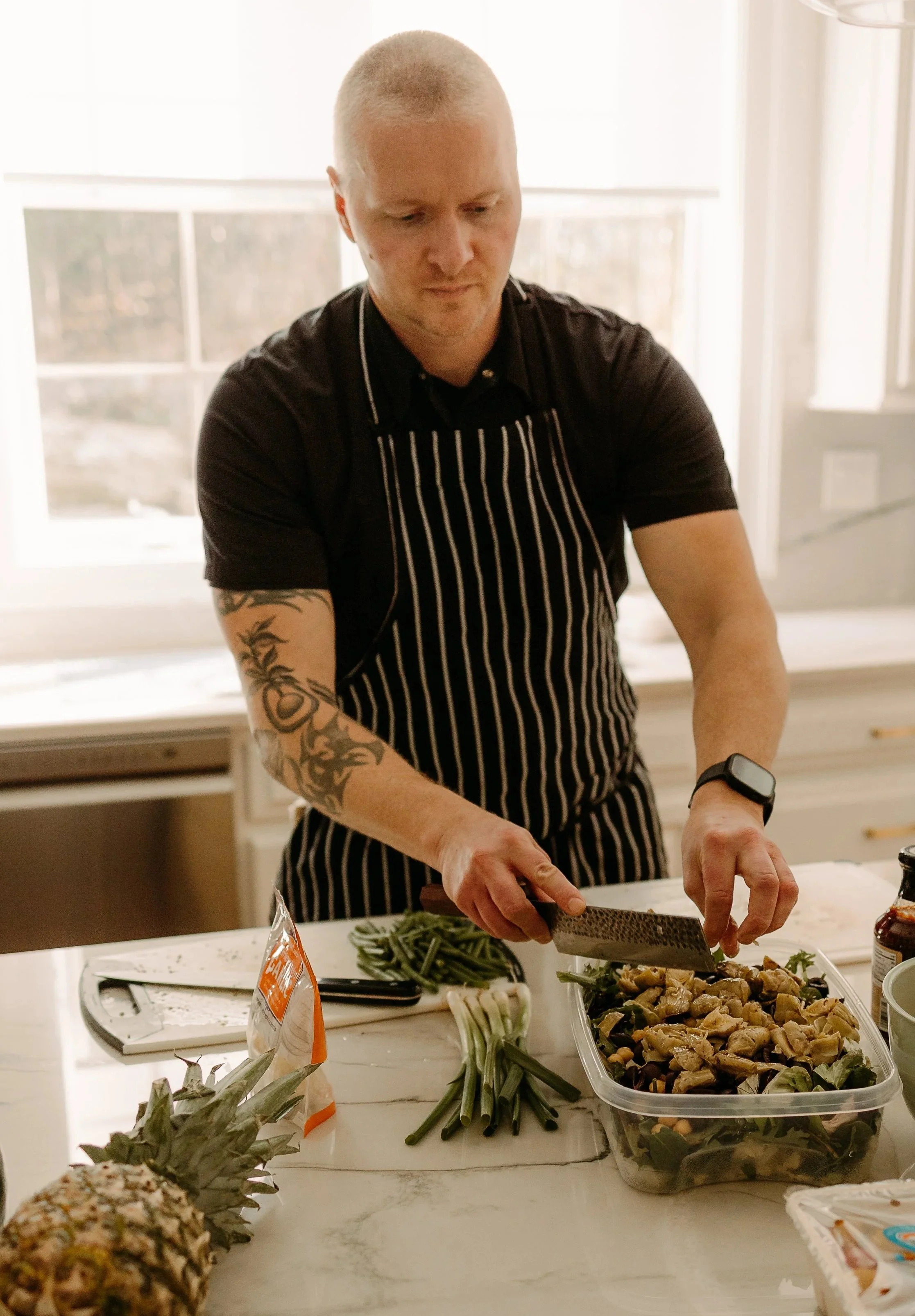 A man with a shaved head and tattoos on his arm is chopping salad in a plastic container in a bright kitchen, wearing a black shirt, striped apron, and a smartwatch.