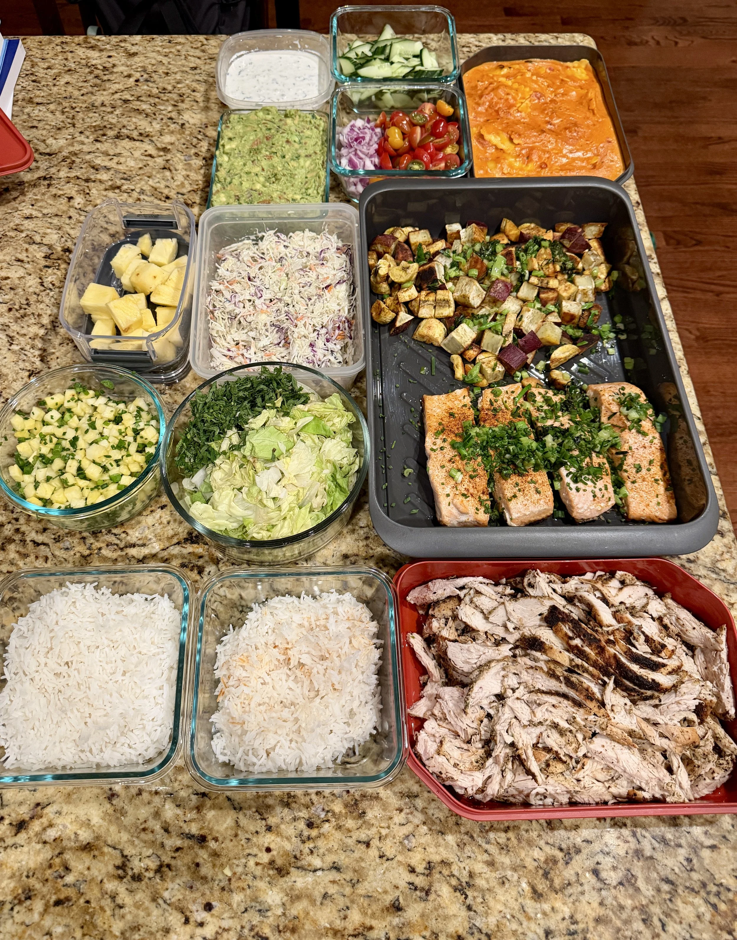 A table filled with various dishes including shredded cheese, cooked chicken, fish fillets garnished with herbs, grilled eggplants, chopped cucumbers, chopped lettuce, coleslaw, pineapple chunks, guacamole, diced pineapple, chopped green herbs, cilan