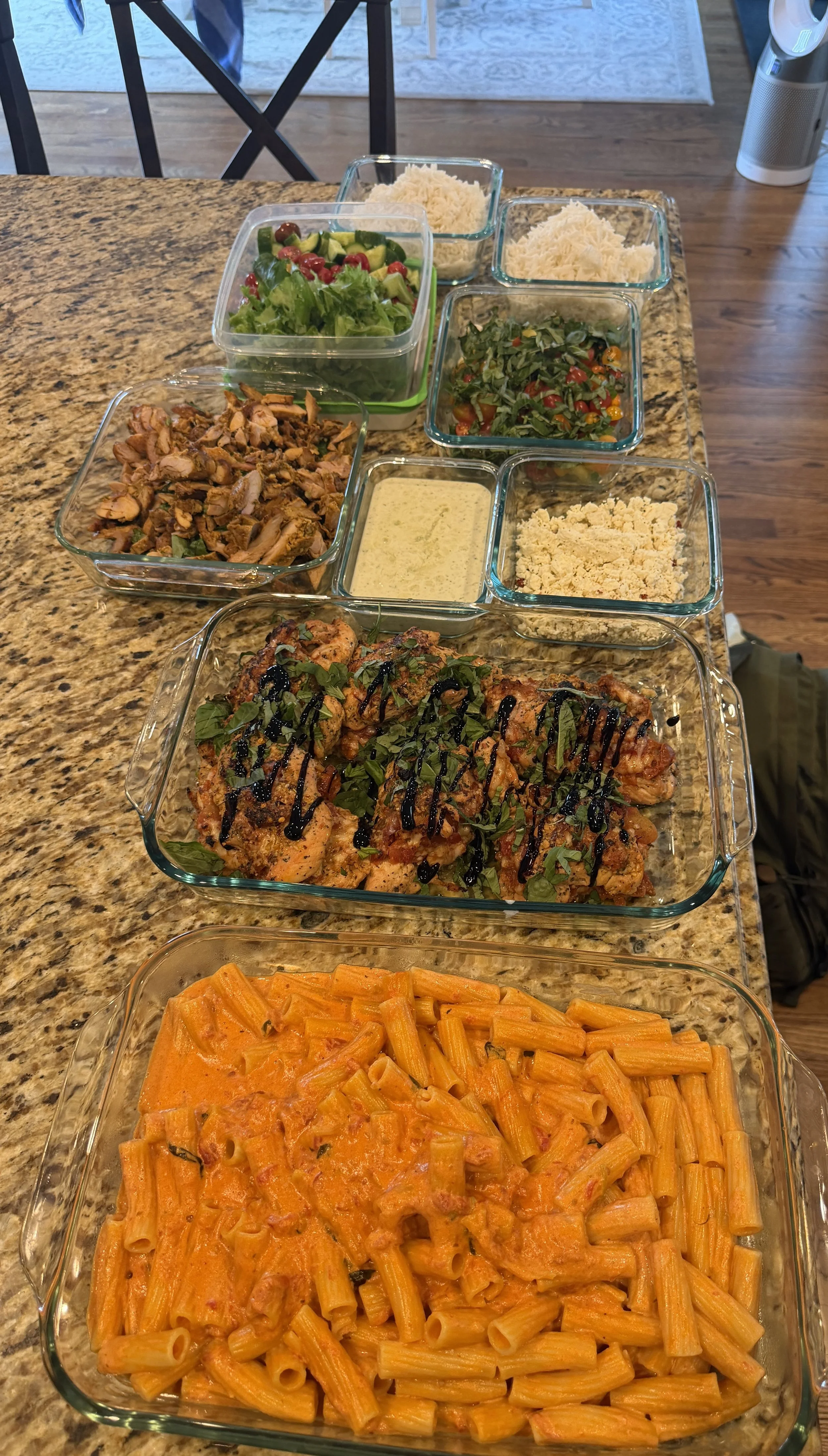 A variety of prepared dishes in glass containers on a kitchen counter, including pasta with tomato sauce, a tray of baked chicken with herbs and balsamic glaze, baked rigatoni with cheese, a vegetable salad, and a green salad with cherry tomatoes and