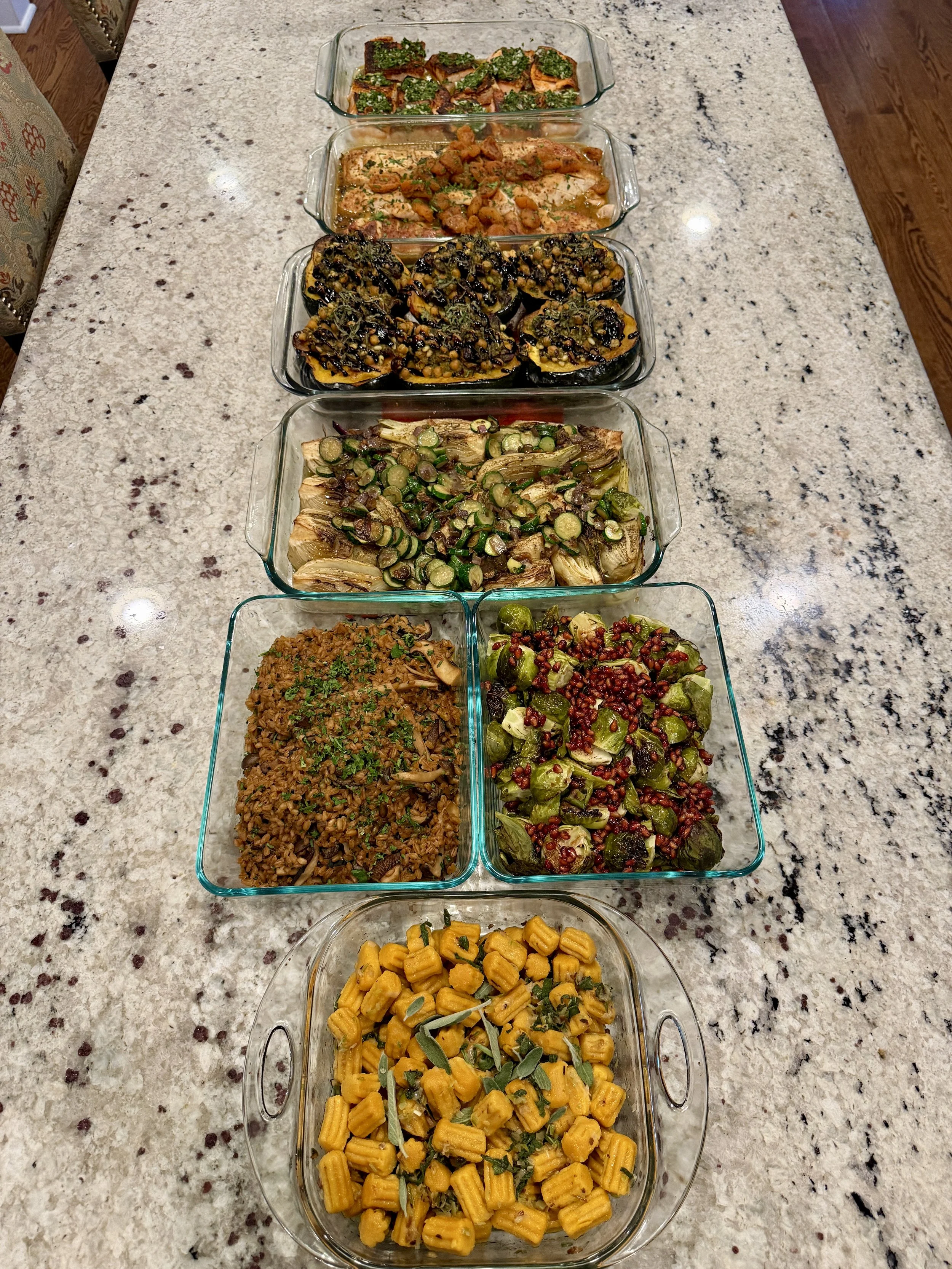 Eight glass dishes filled with various cooked foods arranged on a light-colored granite kitchen countertop.