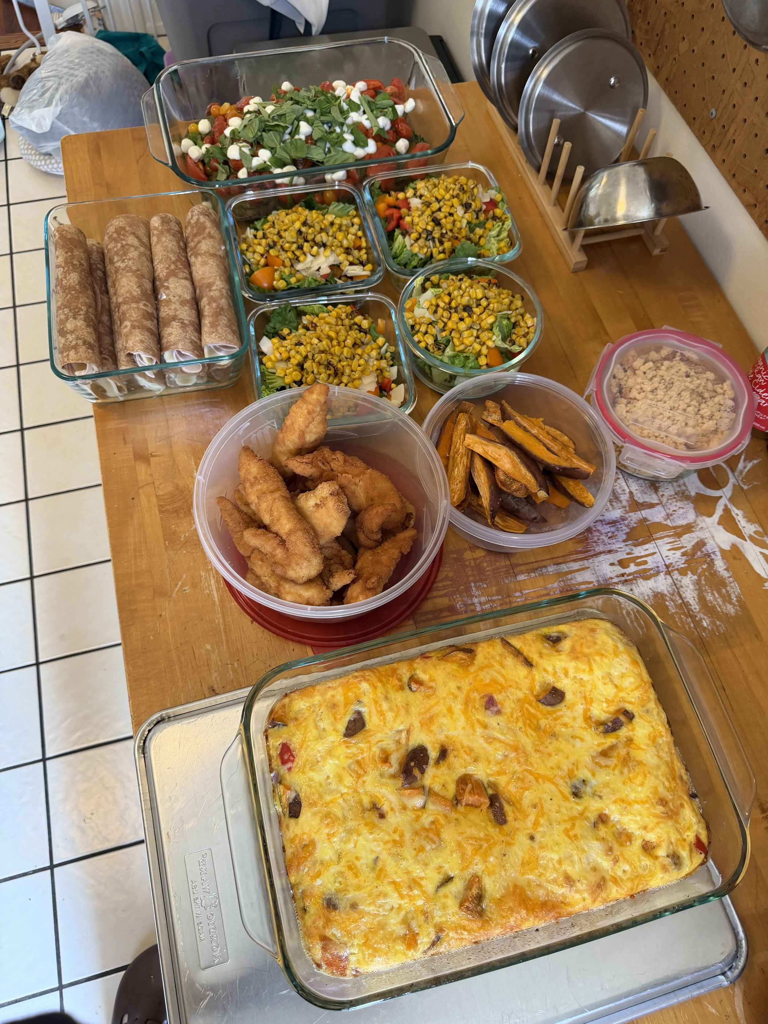 A table with various dishes including a large baked casserole, fried chicken tenders, roasted sweet potatoes, and salads with cherry tomatoes and corn.