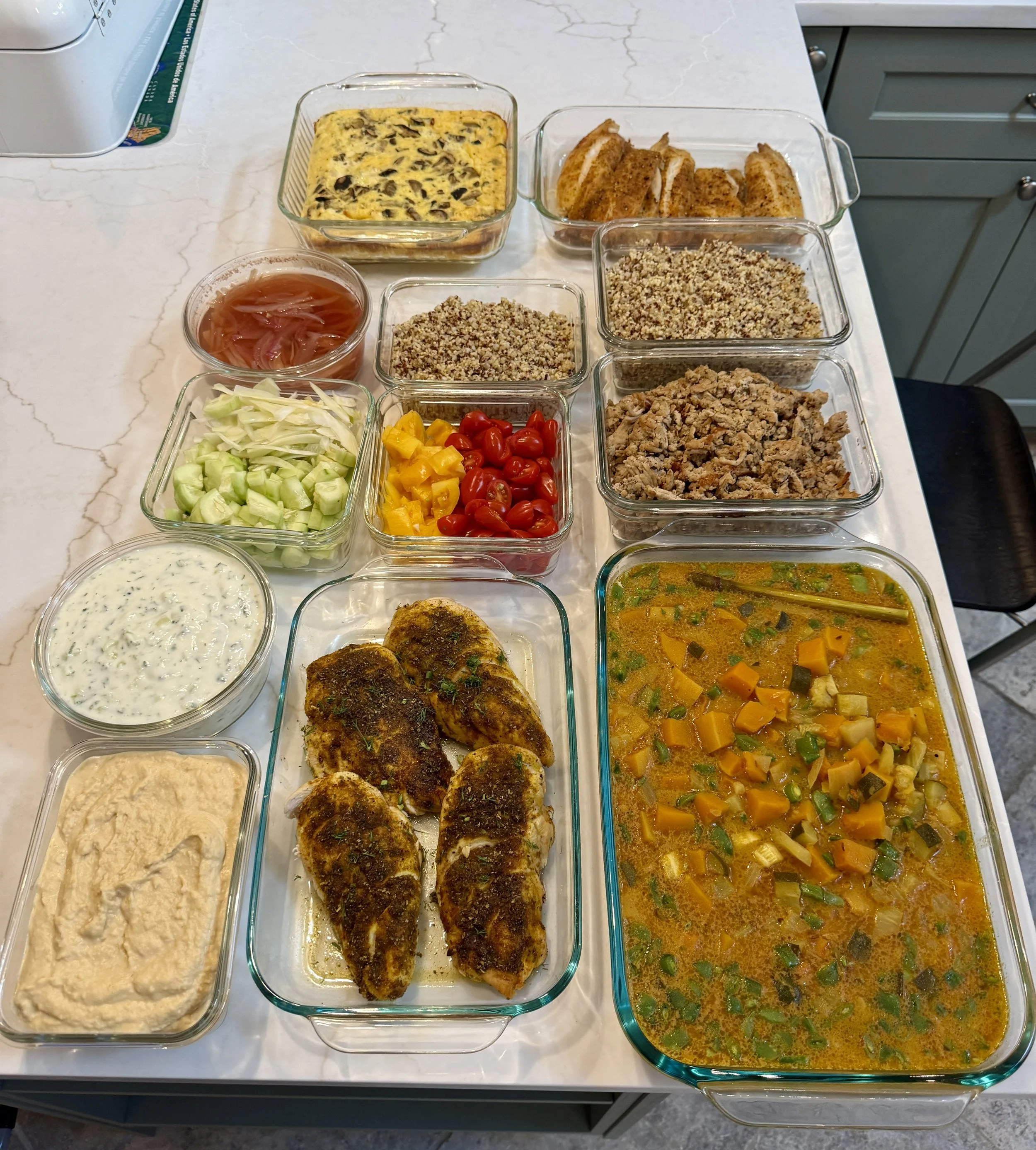 A spread of home-cooked dishes on a kitchen counter, including baked chicken breasts, vegetable soup, rice, various salads, dips, and casseroles in glass containers.