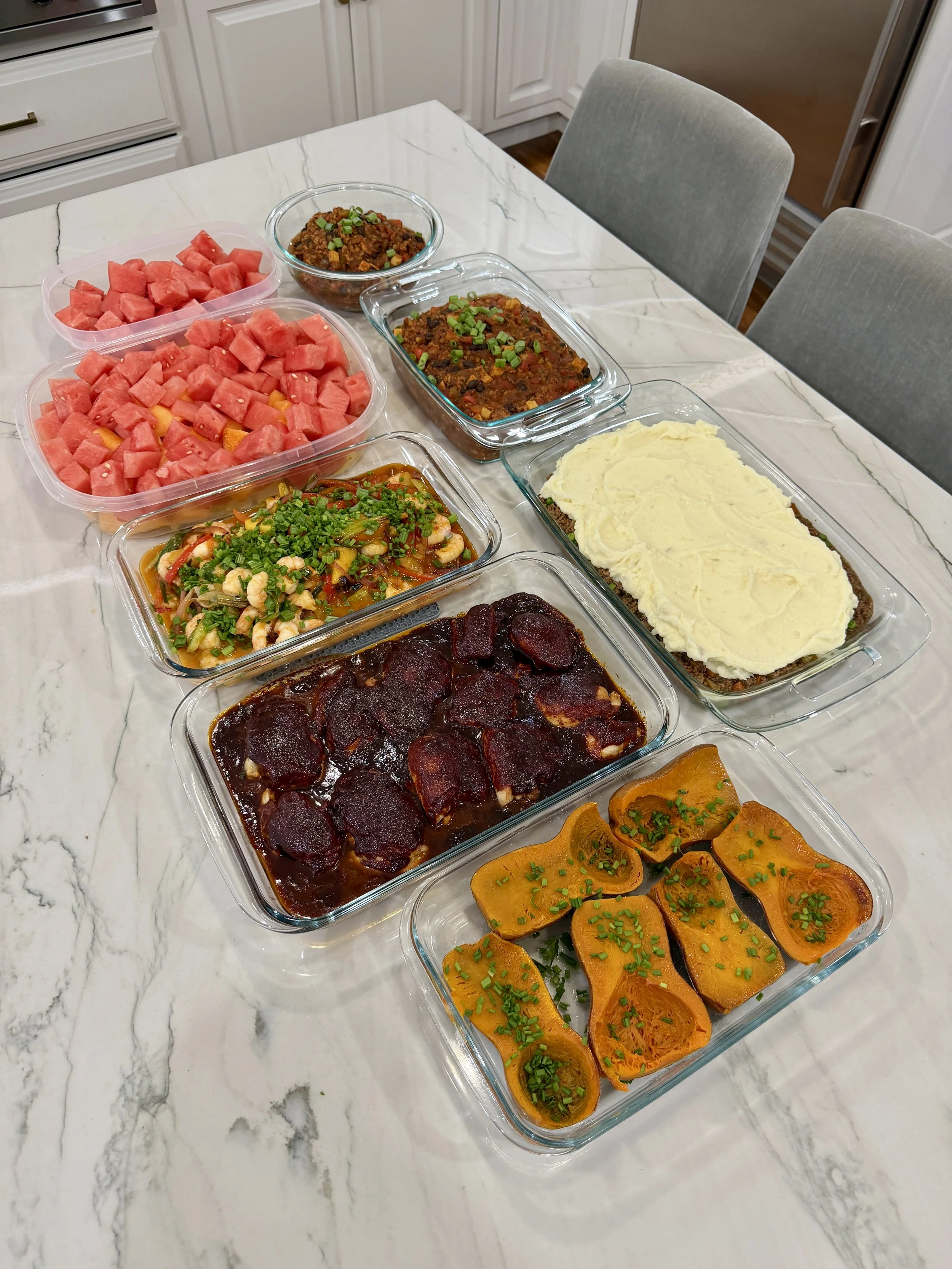 Assorted dishes on a white marble kitchen table, including sliced watermelon, a shrimp dish, a meatloaf with mashed potatoes, beef stew, fried meat patties with sauce, baked squash, chili, and bottled water.