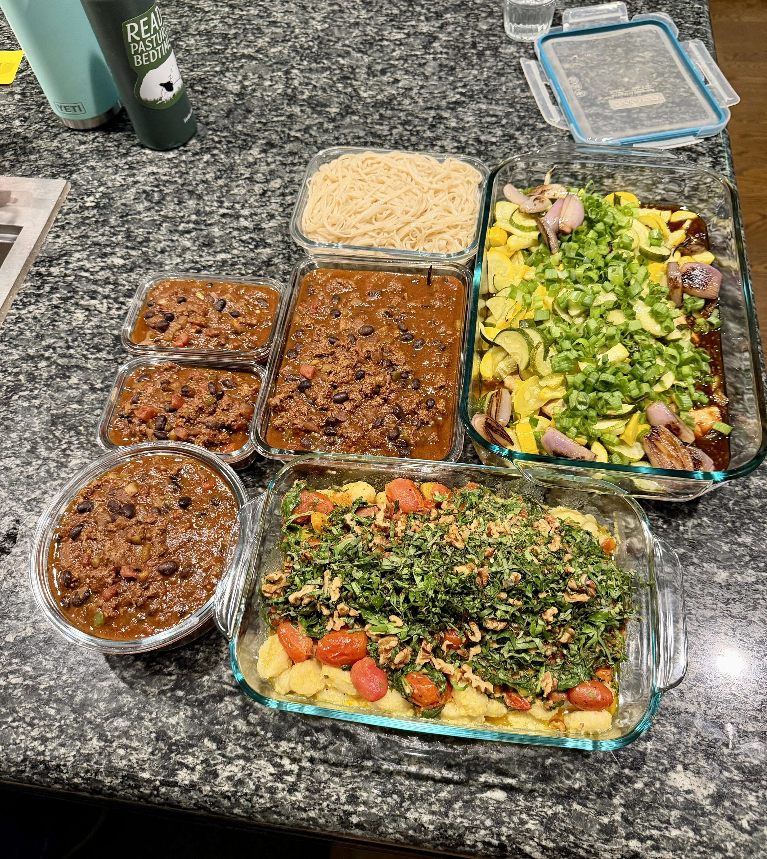 Various cooked dishes in glass containers on a granite countertop, including spaghetti, chili, a vegetable salad with zucchini, yellow squash, onions, and green onions, and a cauliflower and carrot dish.