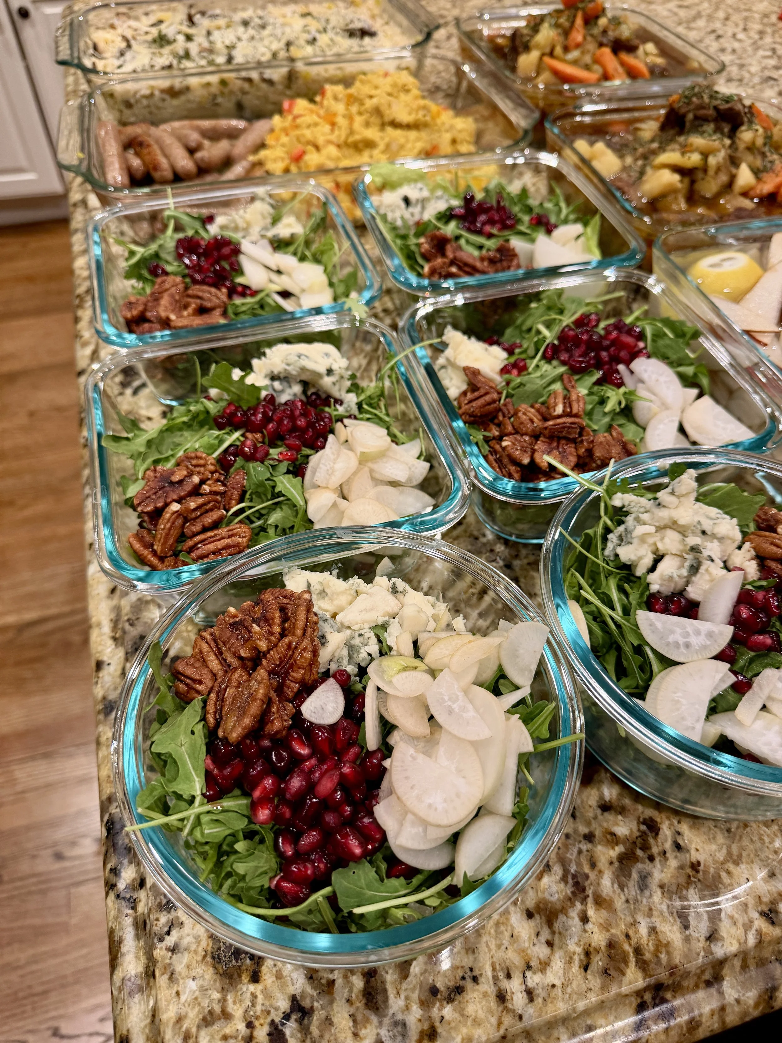 Bowls of fresh salad with arugula, pomegranate seeds, blue cheese crumbles, sliced onions, and pecans on a kitchen countertop, with other food dishes in the background.