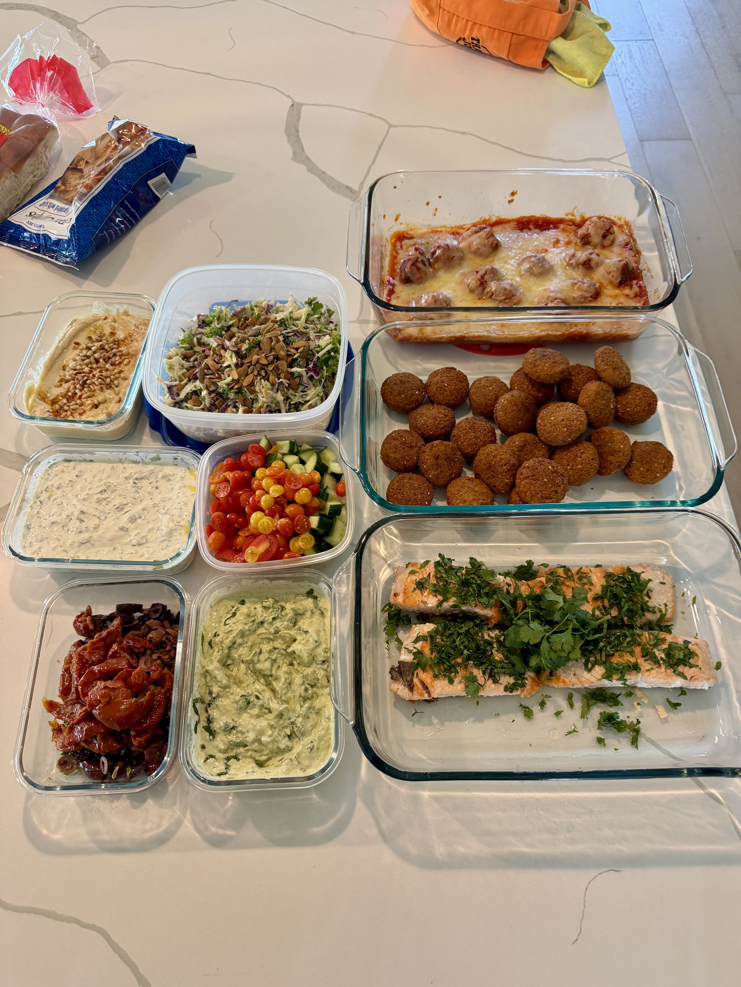 Assorted dishes including baked salmon topped with herbs, falafel balls, various salads, and dips on a white table.
