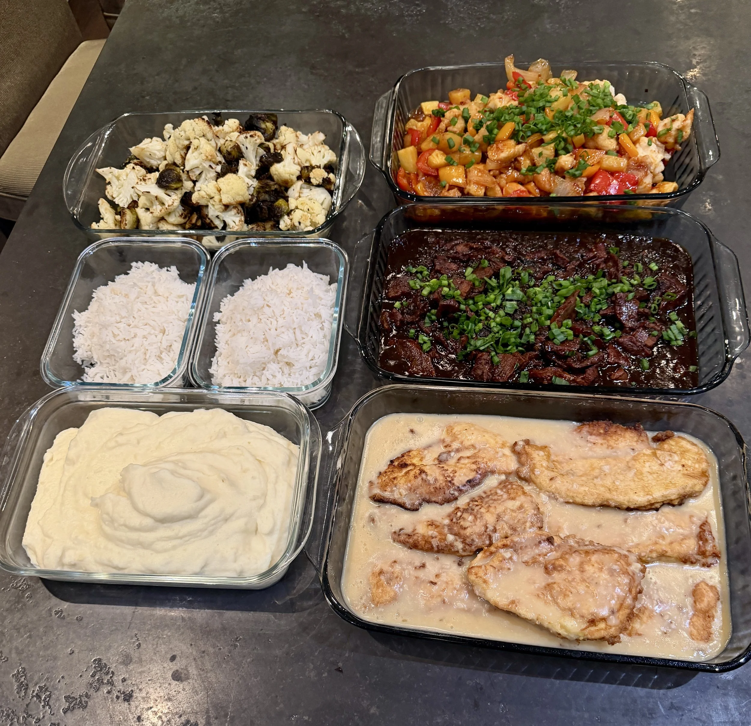 Assorted prepared dishes including mashed potatoes, white rice, a beef stew with green onions, a vegetable stir-fry, roasted chicken in gravy, a cauliflower and mushroom casserole, and a mixed vegetable casserole, all in glass dishes on a dark table.