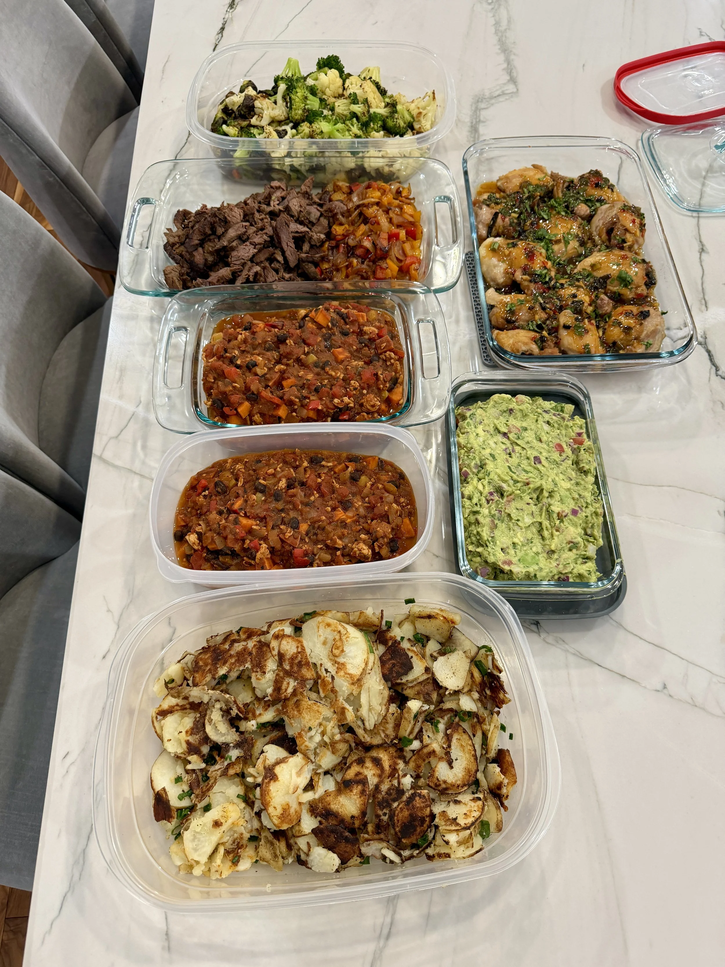 An assortment of cooked dishes in glass and plastic containers on a white marble table, including chopped broccoli, browned beef, chili, taco meat, guacamole, grilled chicken, and sliced cooked potatoes.