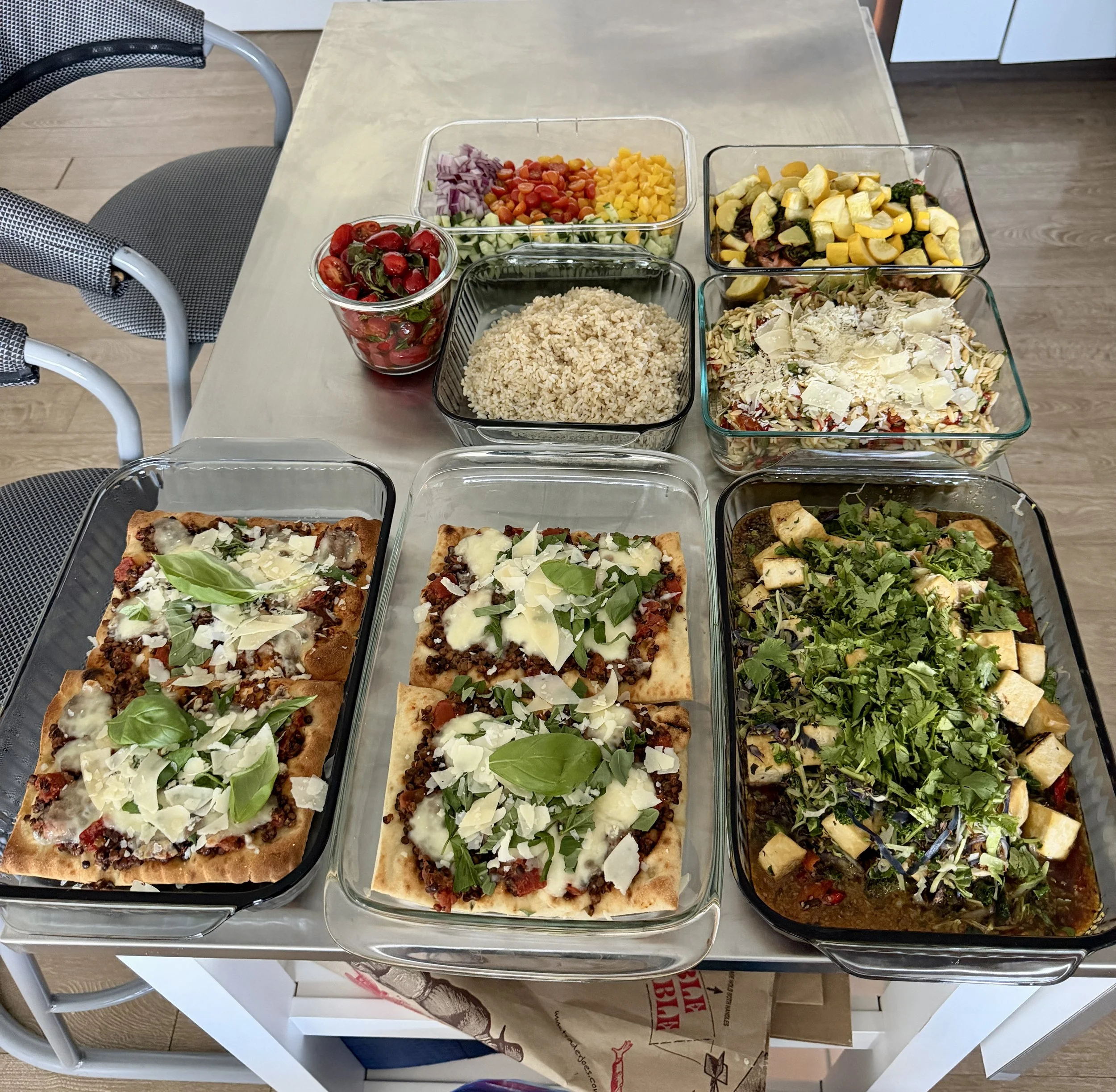 Various salads and toppings in glass containers on a table, including cherry tomatoes, chopped onions, diced yellow bell peppers, cooked rice, chopped zucchini, and a mix of chopped vegetables with cheese, alongside flatbread with toppings.