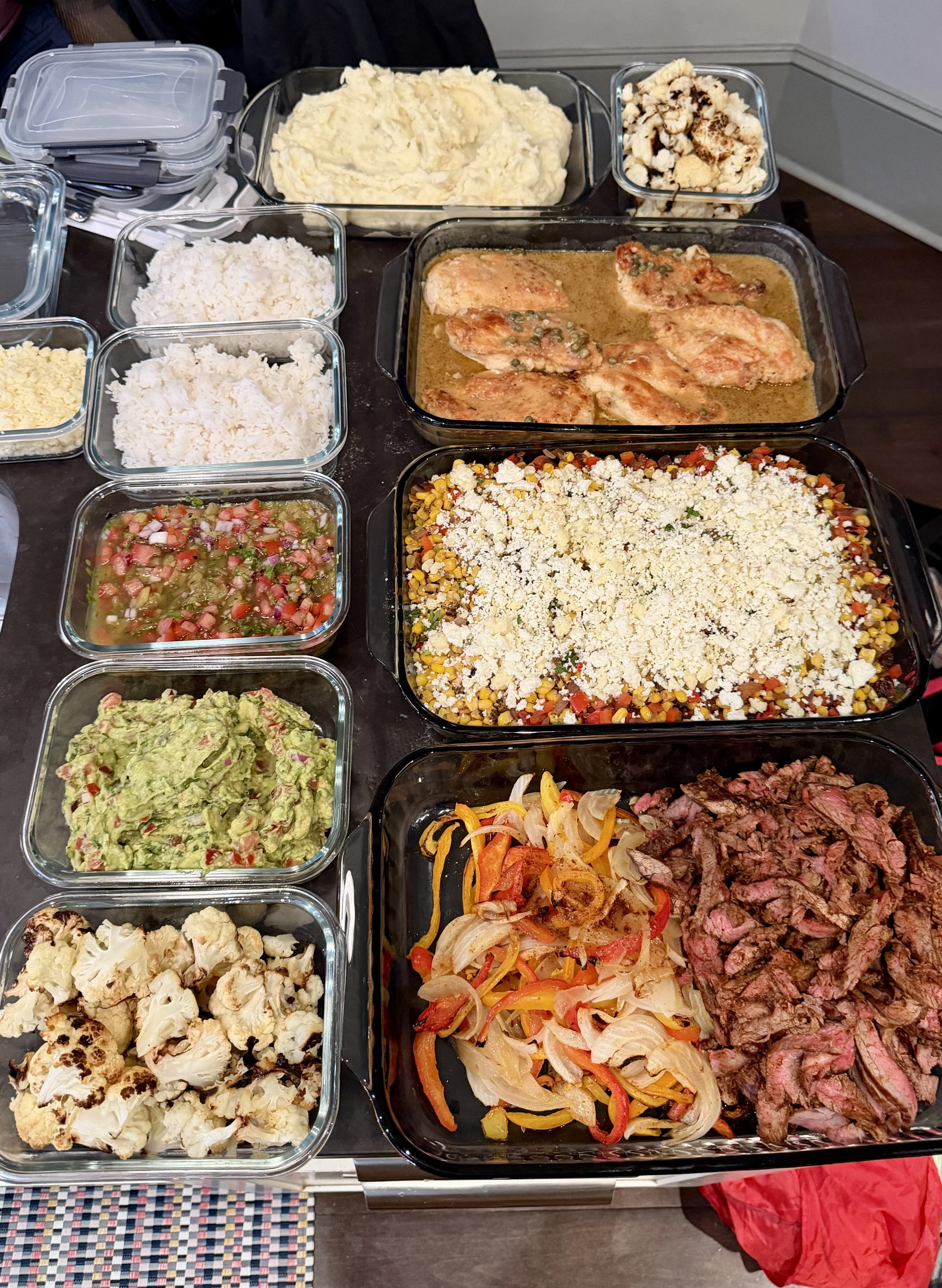 A table filled with various dishes in glass containers, including cauliflower, guacamole, diced tomato salad, cauliflower florets, cooked chicken in sauce, vegetable stir-fry, shredded beef, grilled peppers and onions, rice, and shredded cheese.