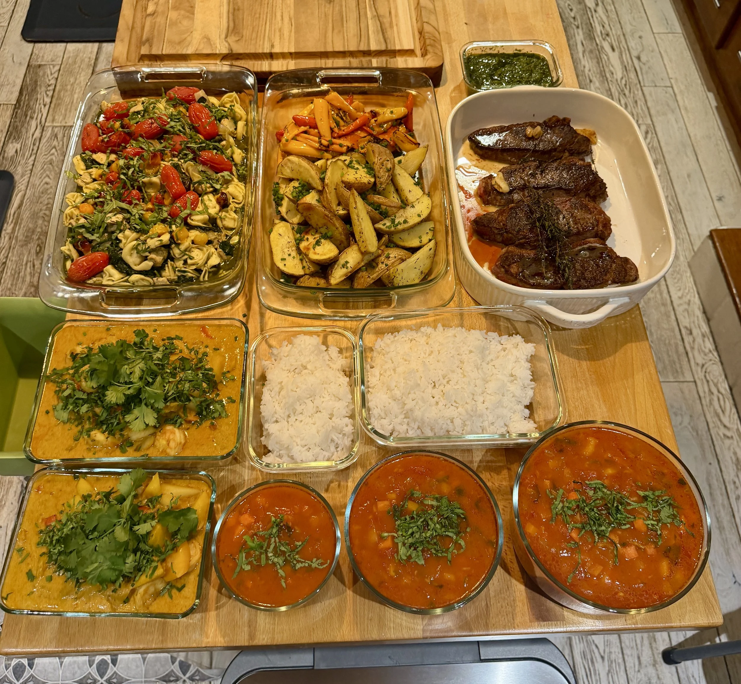 Assorted Indian dishes including vegetable curry, grilled meat, vegetable stir-fry, rice, and three bowls of spicy tomato-based soup, garnished with cilantro on a wooden table.