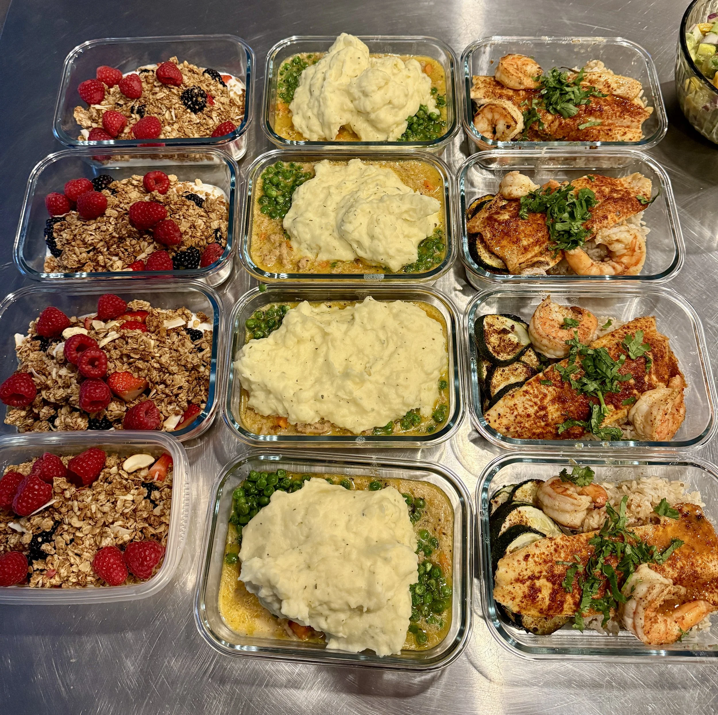 Multiple glass containers filled with food and desserts on a stainless steel surface. The containers include fruit granola parfaits with raspberries and blackberries, mashed potatoes with green peas and carrots, and baked fish with zucchini and shrim