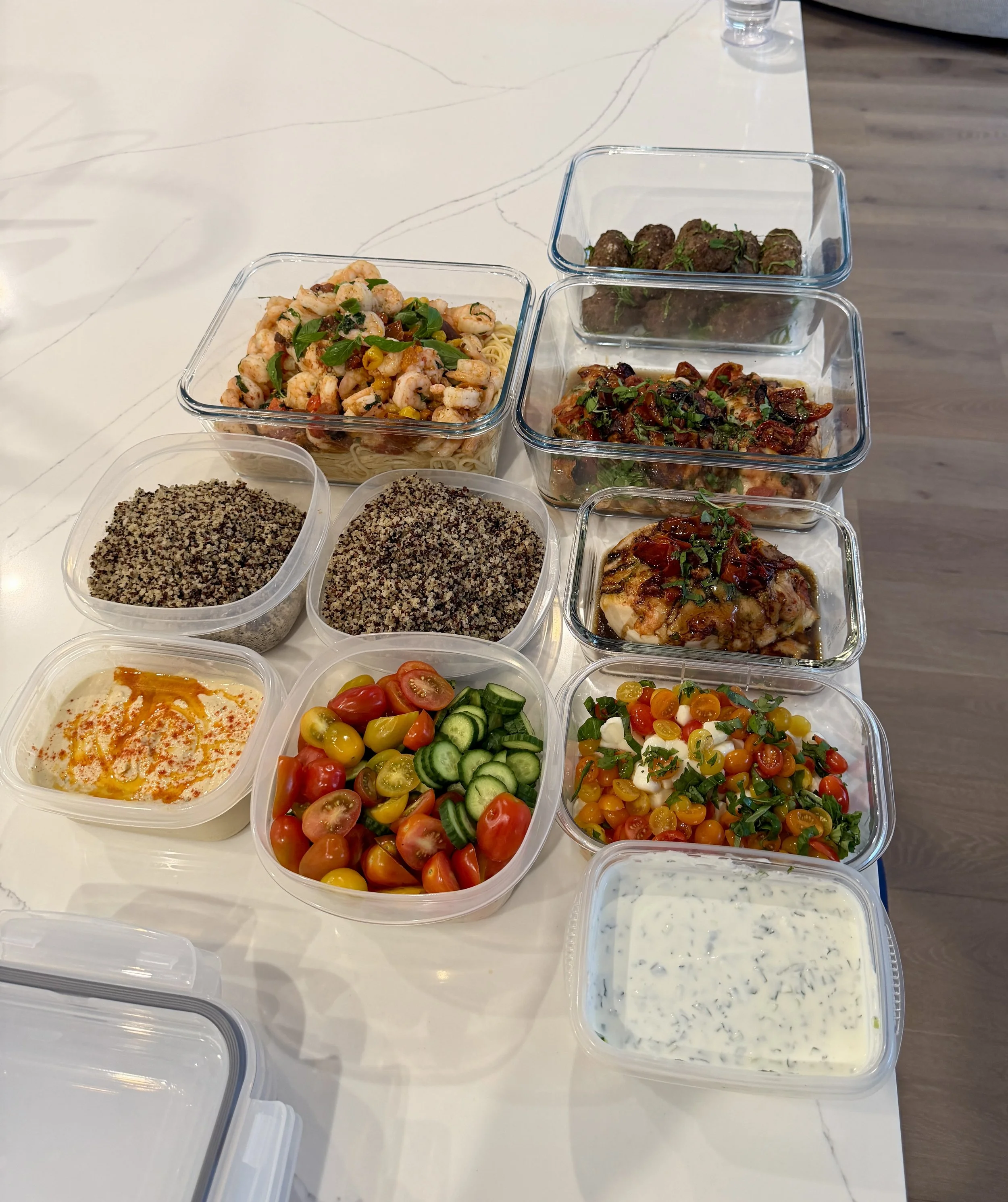 Assorted dishes in clear plastic containers on a white table, including shrimp and pasta, meatballs, chicken with sauce, chopped vegetables, cherry tomatoes, and creamy herb dressing.