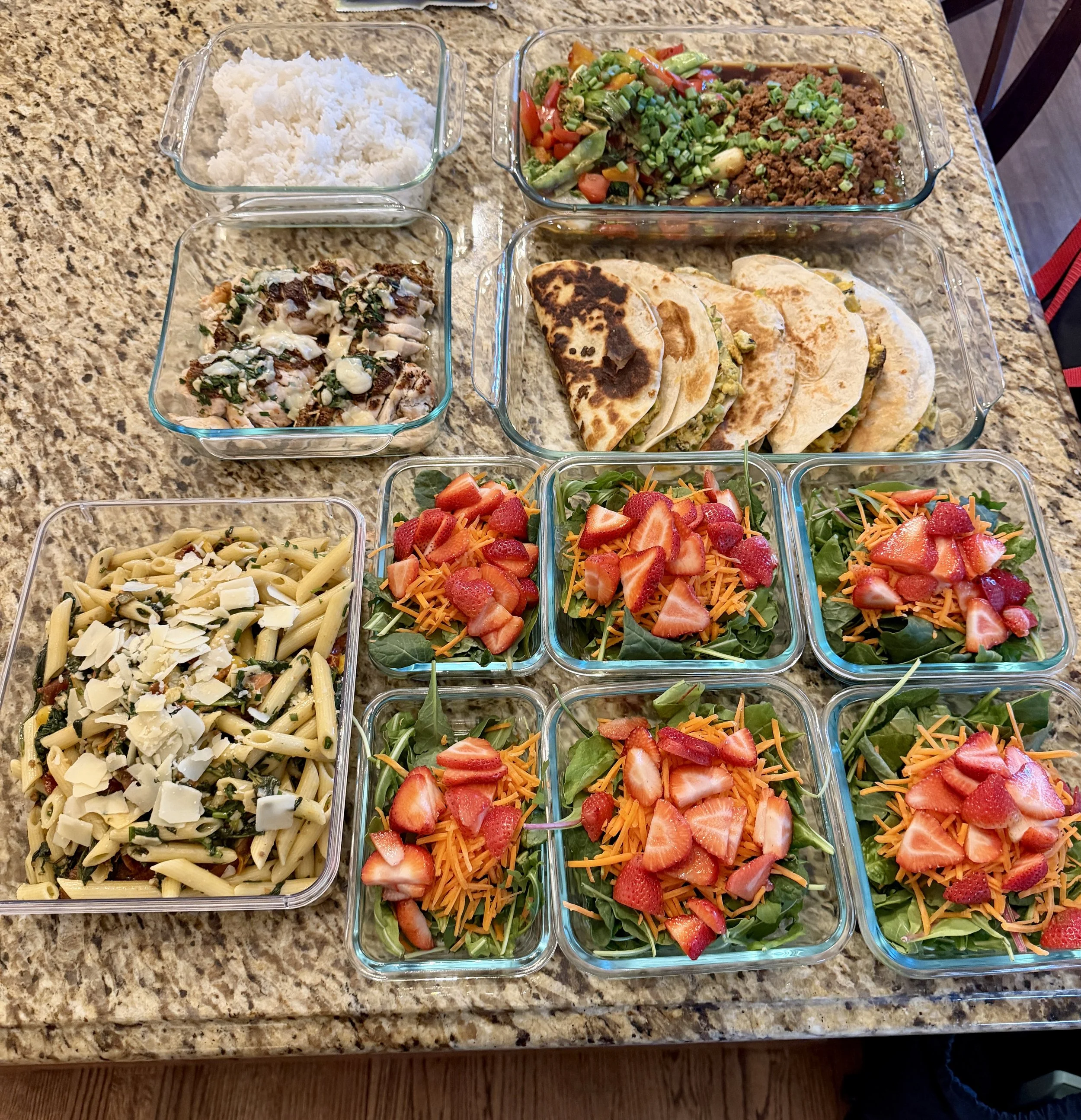 An assortment of prepared food items on a kitchen countertop, including pasta salad, chopped vegetable salad, shredded chicken, rice, tacos filled with meat and vegetables, and strawberry salads with shredded carrots, spinach, and cheese.