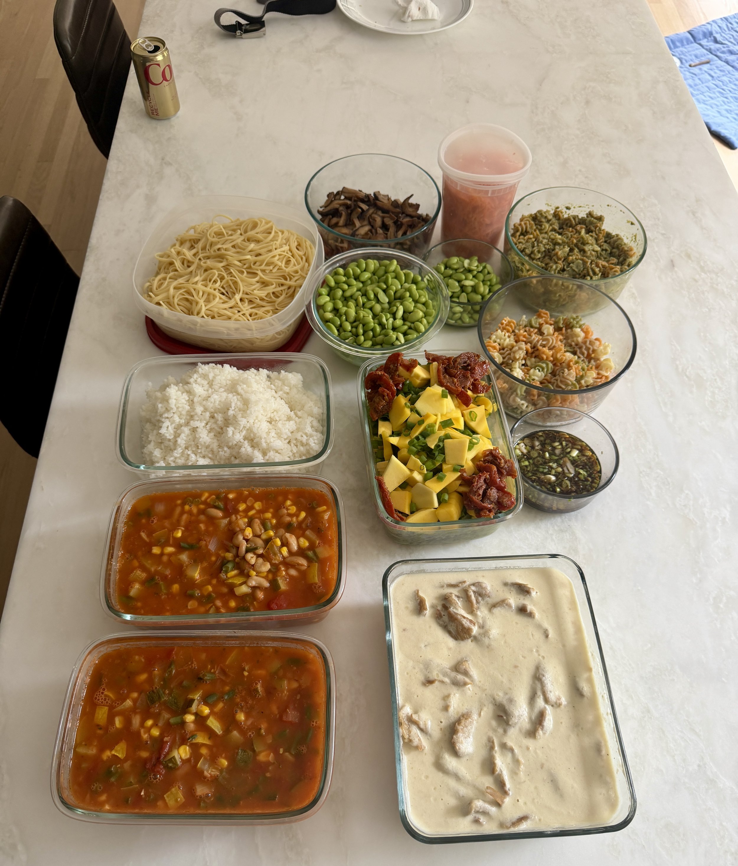 A variety of food items on a kitchen countertop, including bowls of pasta salad, green beans, lasagna, and a container of soup, with a can of soda and some other items in the background.