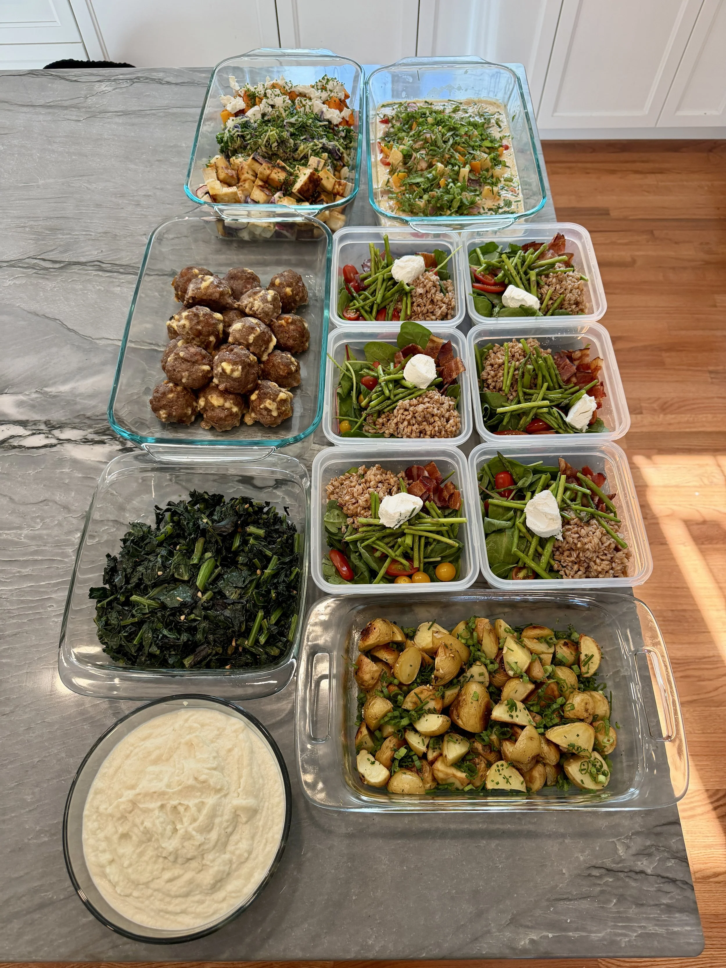 Assorted prepared salads and dishes in glass and plastic containers on a kitchen counter, including greens, roasted potatoes, meatballs, and a creamy dip.