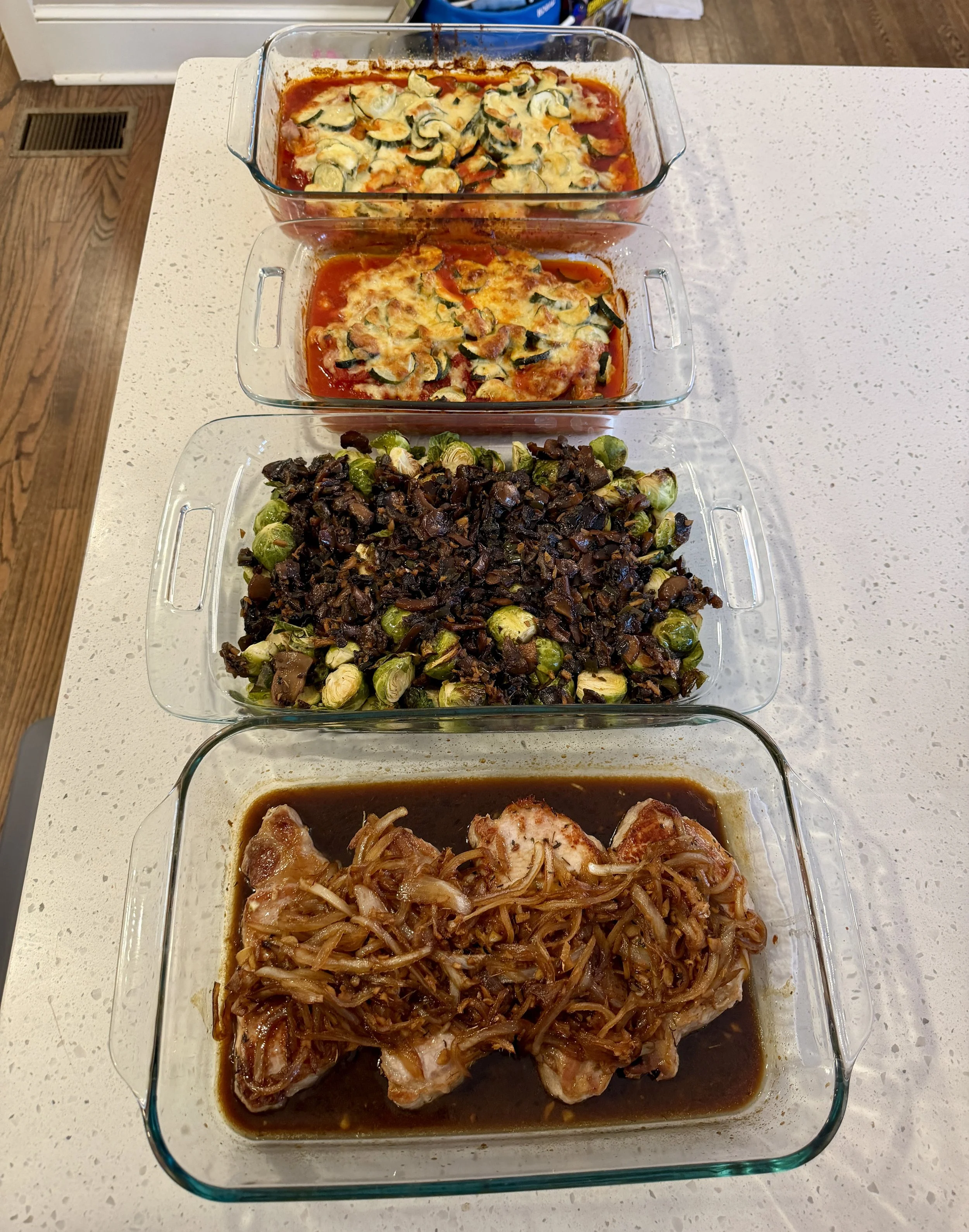 Four glass baking dishes filled with different cooked dishes on a white countertop. The first dish contains baked zucchini casserole, the second contains baked chicken parmesan, the third has sautéed Brussels sprouts with chopped dark vegetables, and