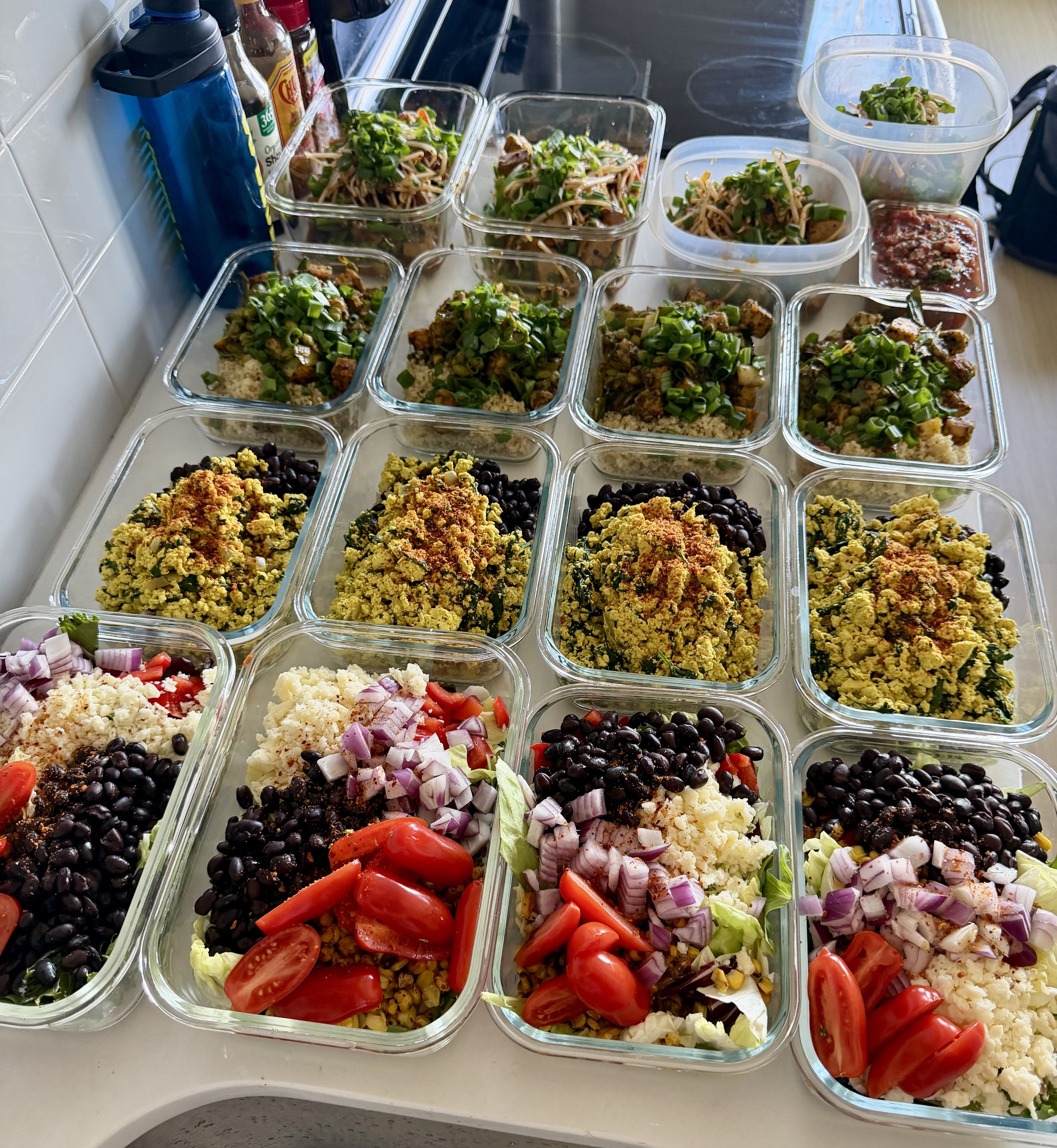 A table filled with several glass containers of different salads, including bean salads, couscous salads, and vegetable salads like cherry tomatoes, onions, and lettuce, prepared for a gathering or meal.