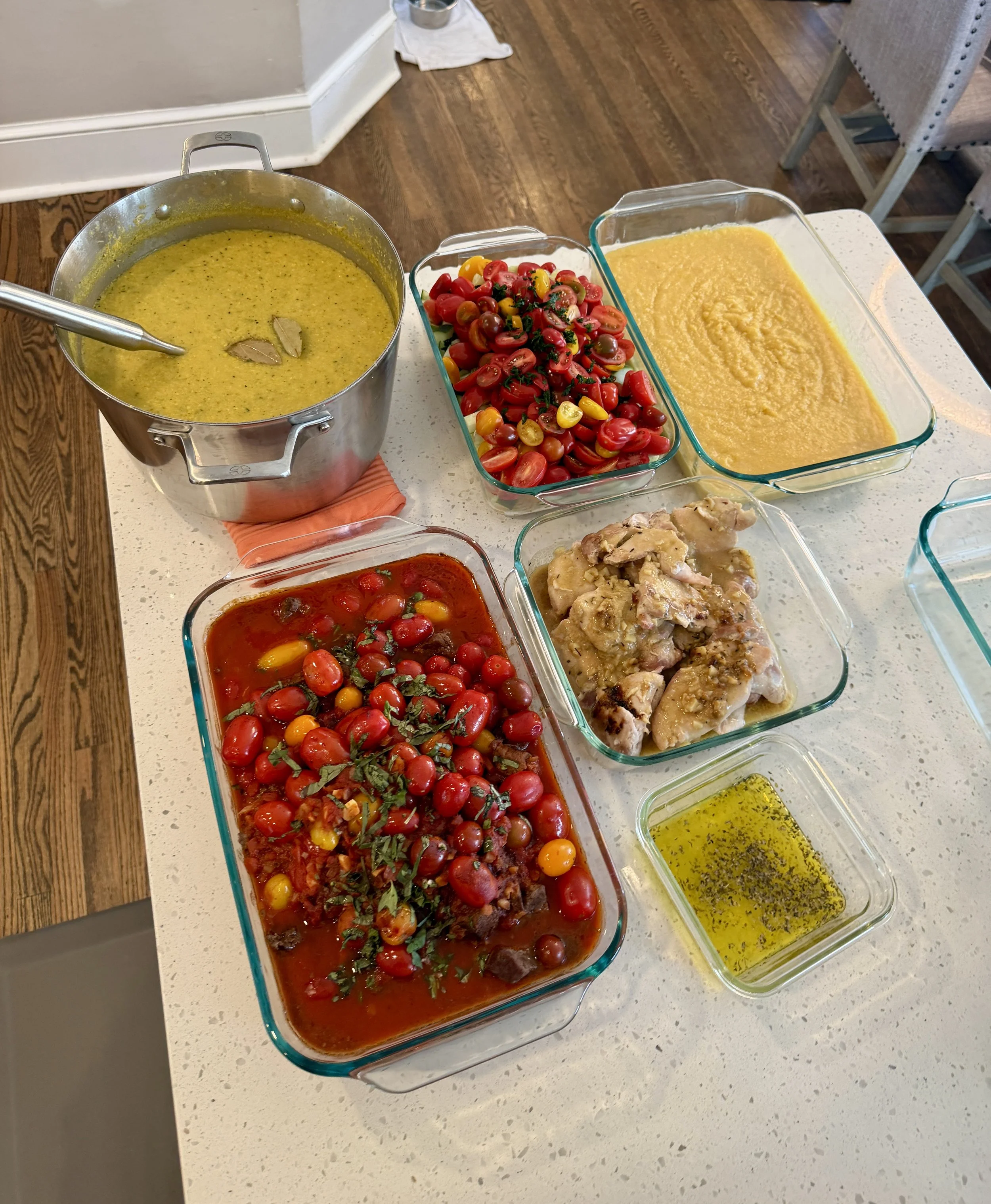 A collection of food items on a kitchen counter including a pot of yellow soup, a glass dish of cherry tomatoes, a dish of yellow pudding or custard, and a dish of cooked chicken.