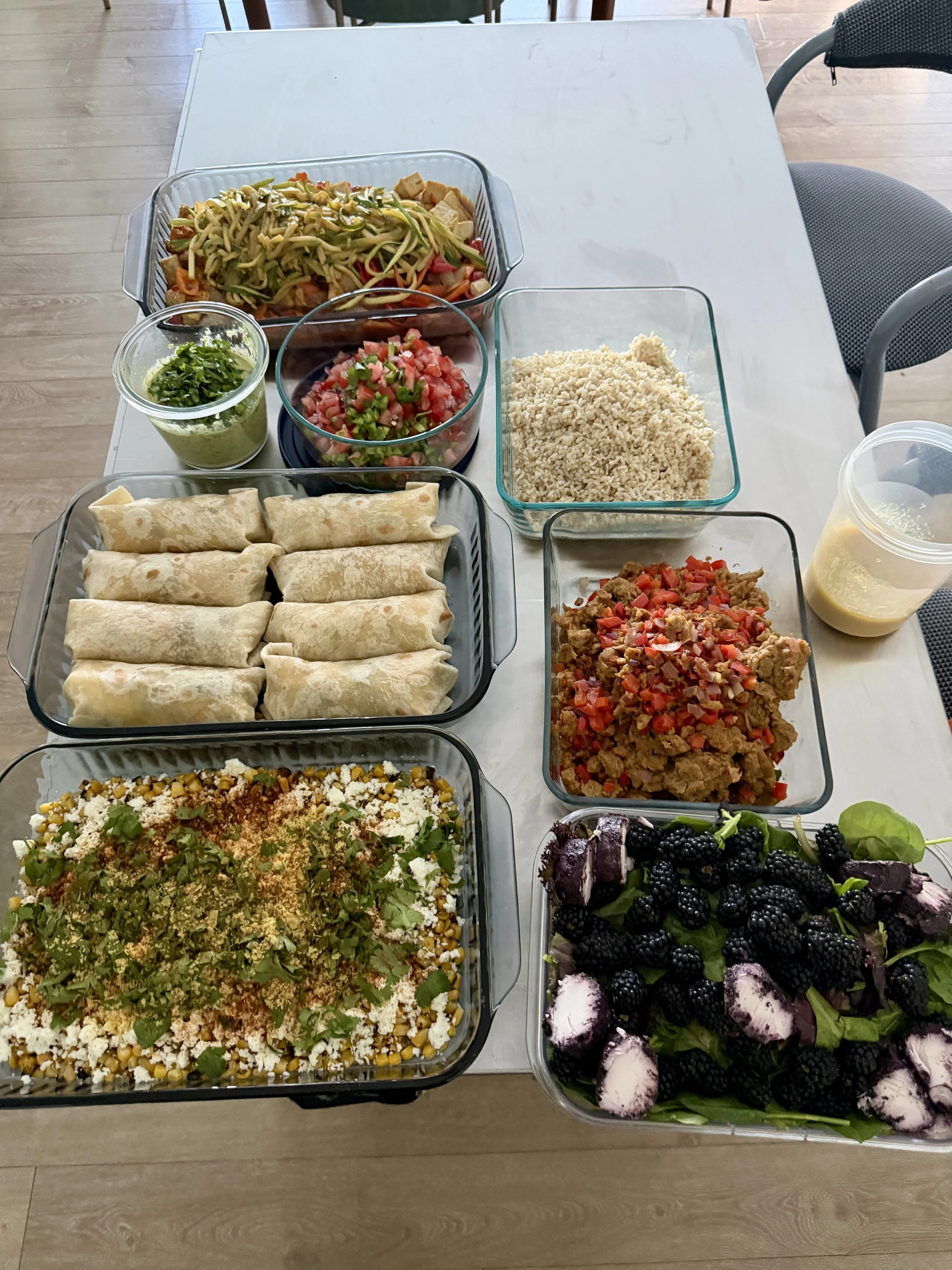 A table with various dishes including a large bowl of salad topped with herbs, a plate of rolled tortillas, a bowl of chopped tomatoes and herbs, a bowl of cooked rice, a bowl of ground beef with vegetables, a small container of guacamole, a large sa