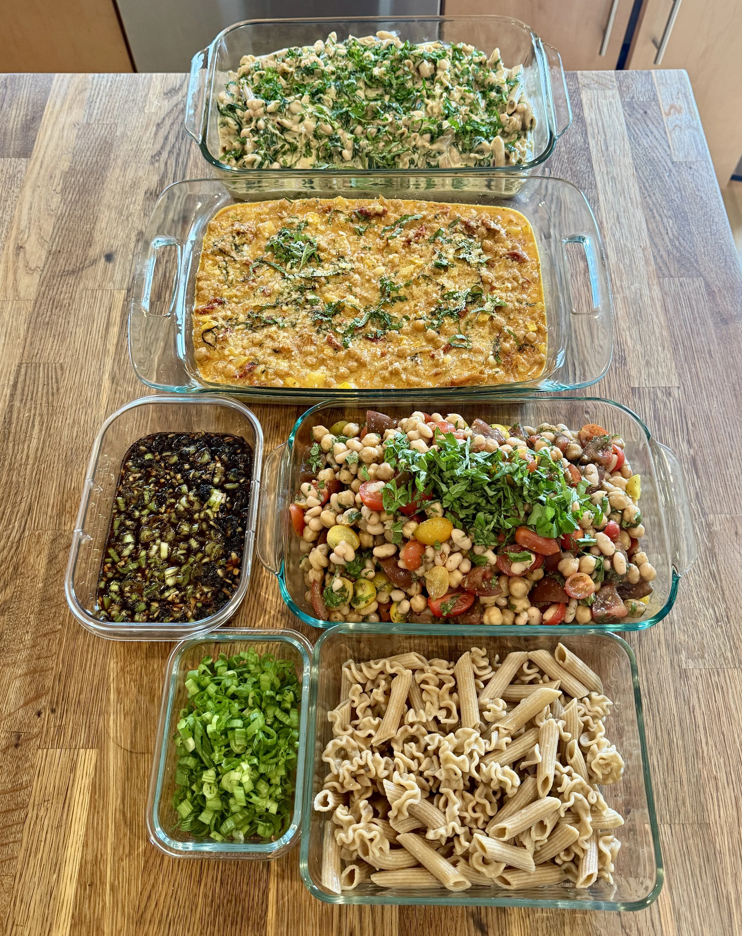Assorted cooked vegetarian dishes in glass baking dishes on a wooden table, including pasta salad, bean salad, vegetable chili, hummus, and a mixed vegetable medley garnished with herbs.