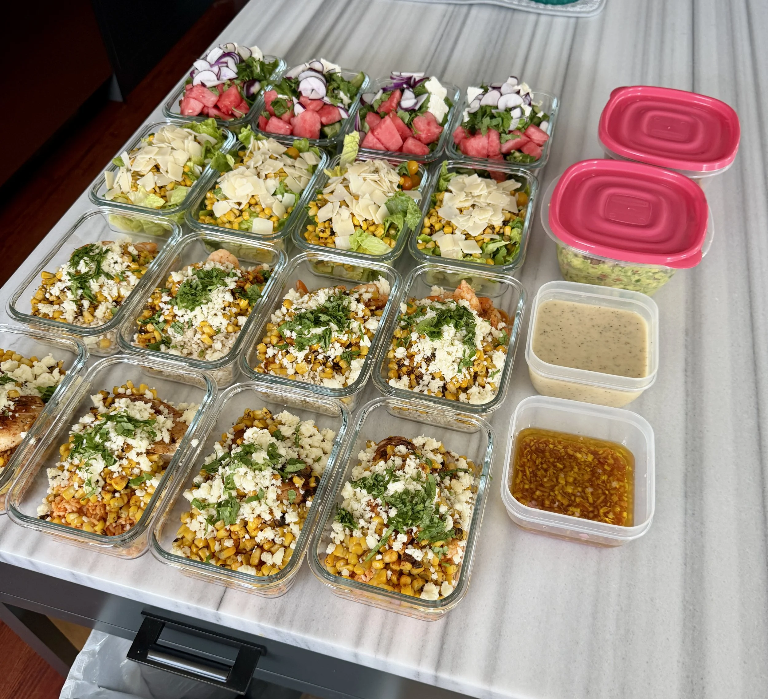A table with multiple glass containers filled with various salads, including corn, cheese, vegetables, and spinach, along with two empty plastic containers with pink lids and a small bowl of dressing.