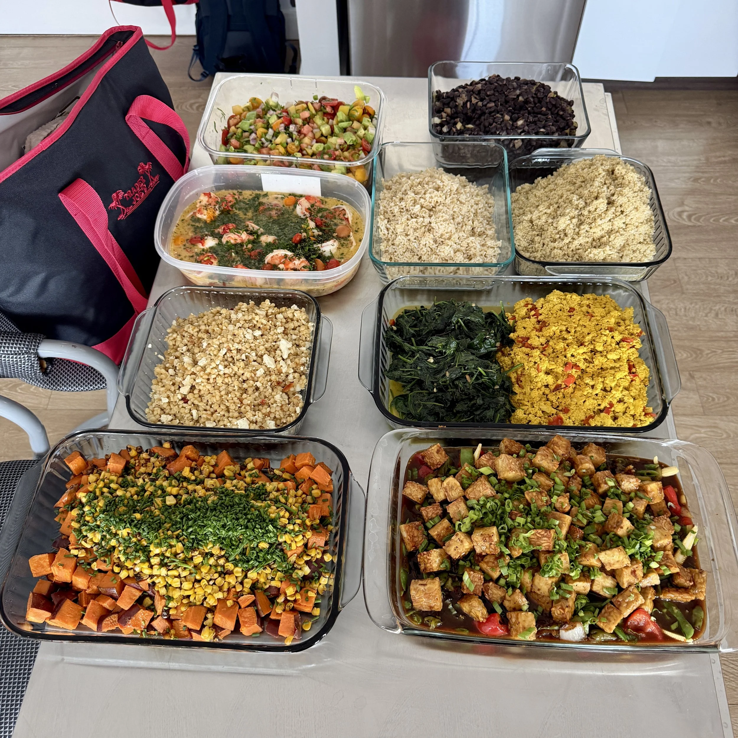 A table filled with various prepared dishes in glass containers, including salads, grains, cooked vegetables, and tofu, with a black and pink bag on the side.