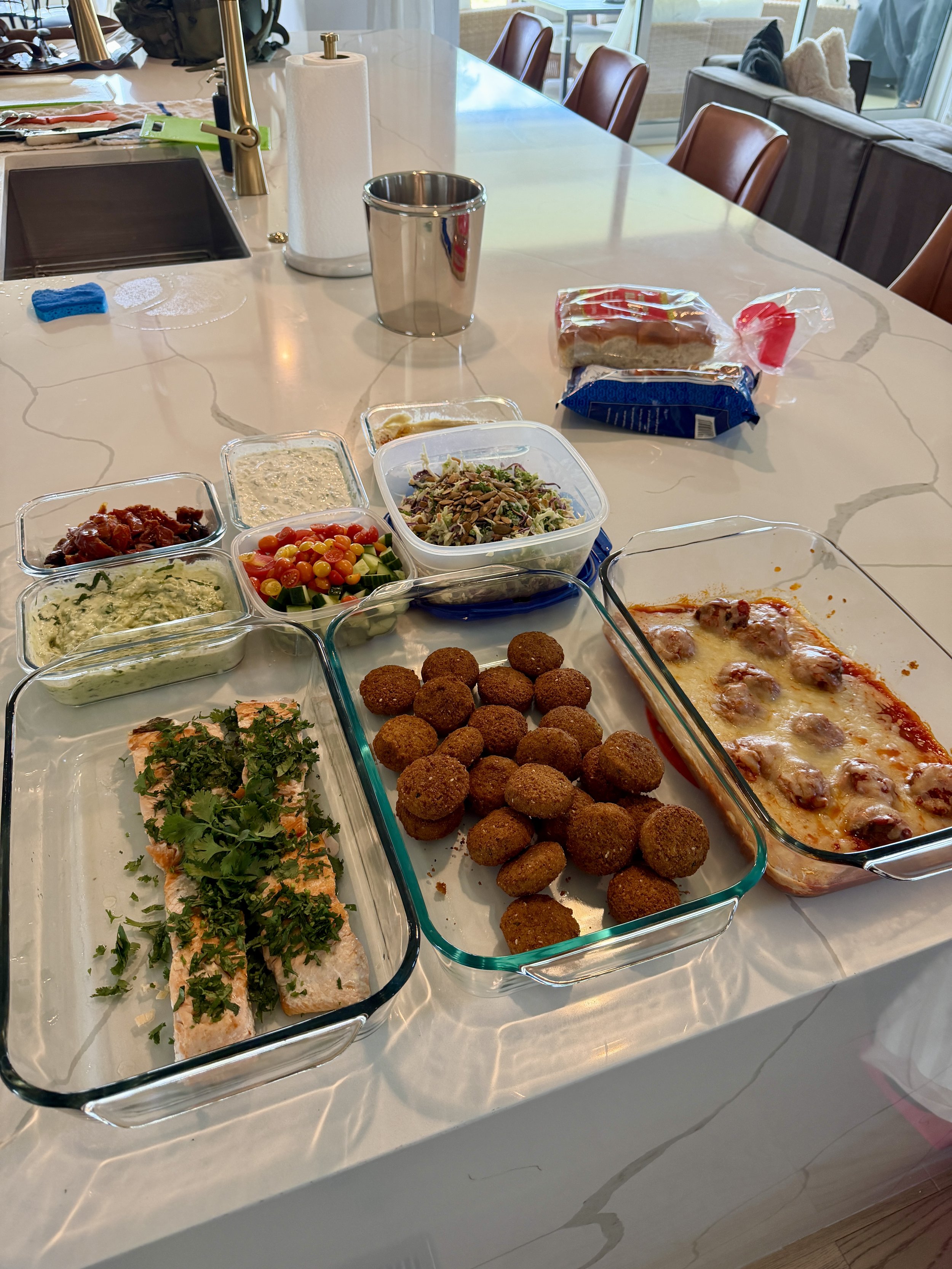 A kitchen island with various dishes including salad, dip, croquettes, salmon, pizza, and bread in glass containers and plates on a white marble countertop.