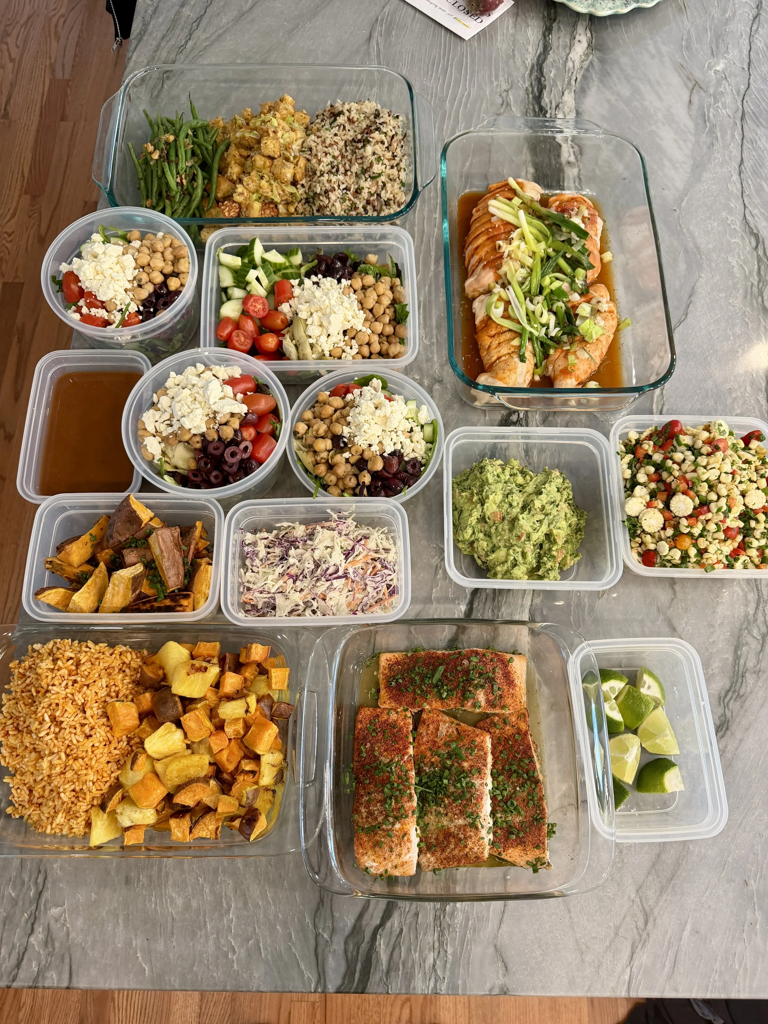 Assorted prepared home-cooked dishes on a marble countertop, including salmon fillets, rice, roasted vegetables, salads with chickpeas, cherry tomatoes, and feta, guacamole, lime wedges, and green beans.