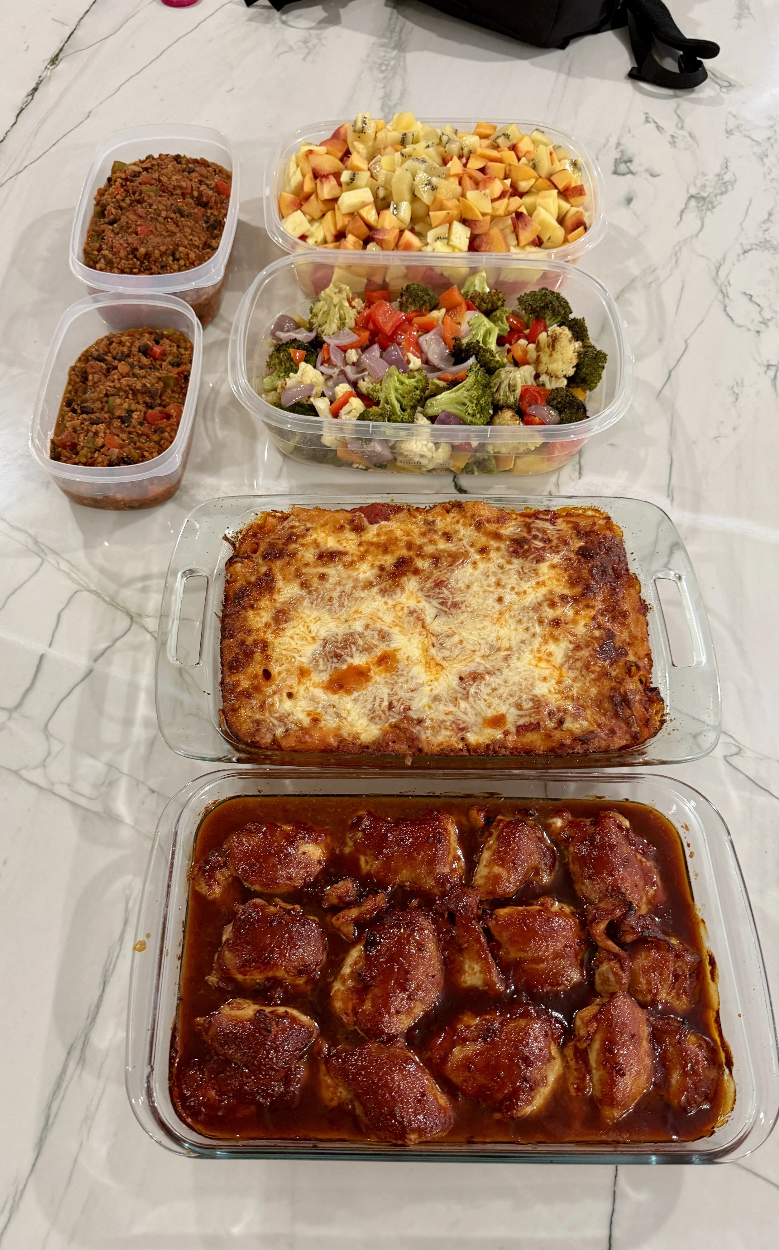 A spread of homemade foods on a marble countertop, including a baked dish with cheese, a bowl of barbecue chicken, a salad with broccoli, carrots, cauliflower, and bell peppers, two containers of ground beef chili, and two small containers of chili c
