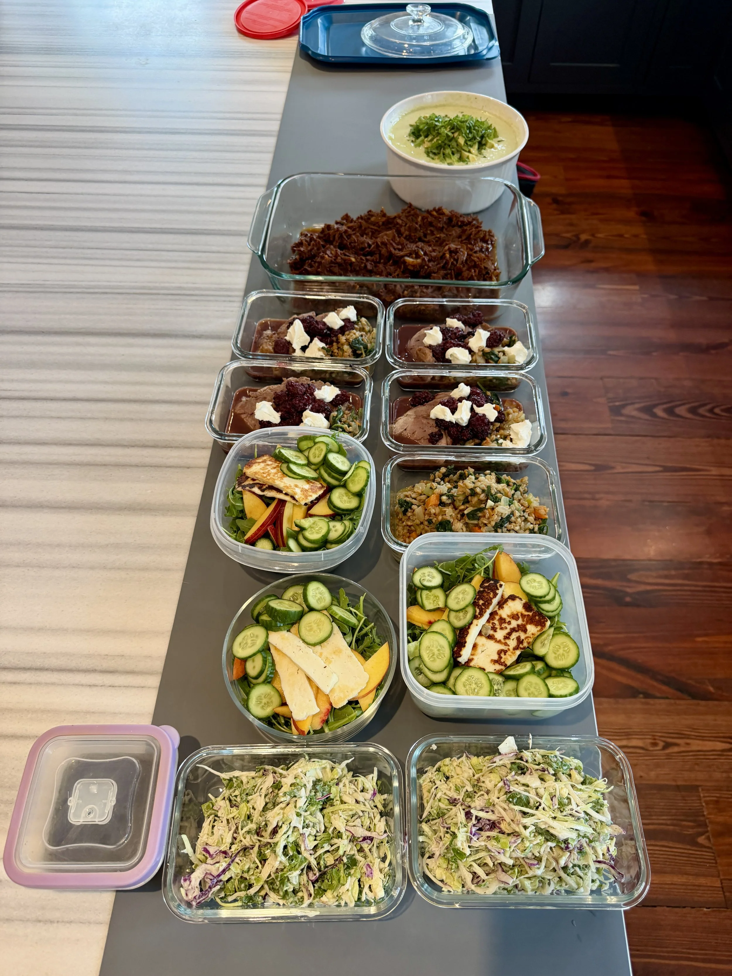 A variety of prepared foods in glass containers on a table, including salads with cucumbers and slices of grilled chicken, bowls of shredded meat, and other dishes with toppings like cranberries, cheese, and herbs.