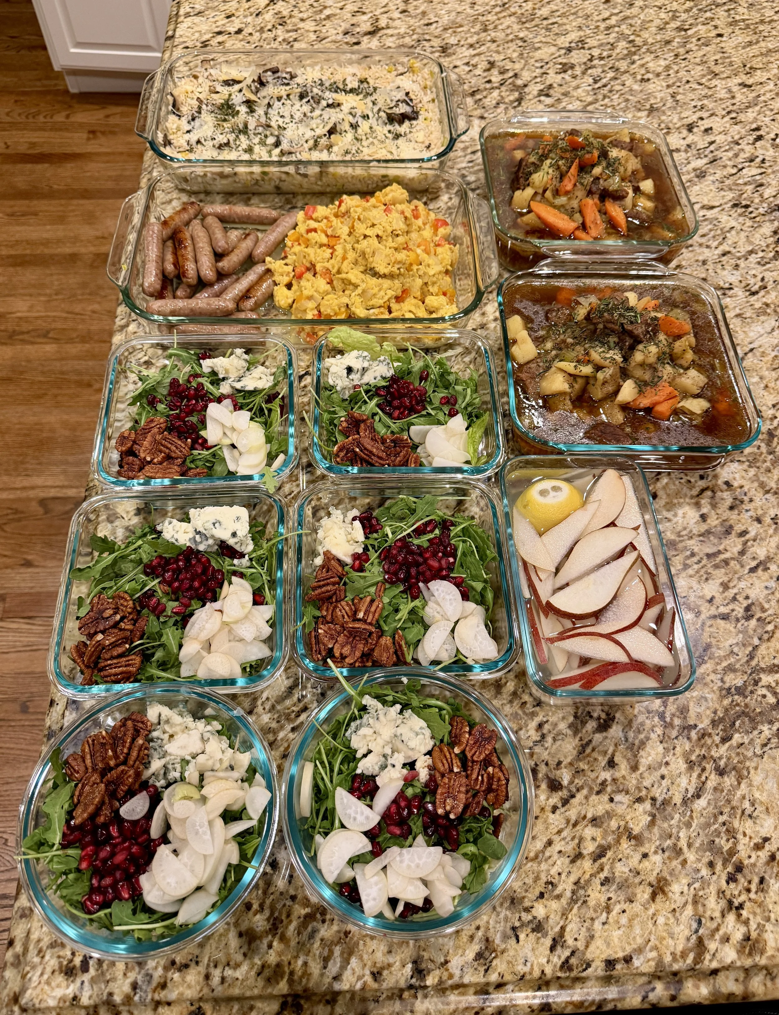 Assorted prepared dishes, including salads, roasted vegetables, mashed potatoes, and cooked sausages, arranged in glass dishes on a granite kitchen countertop.