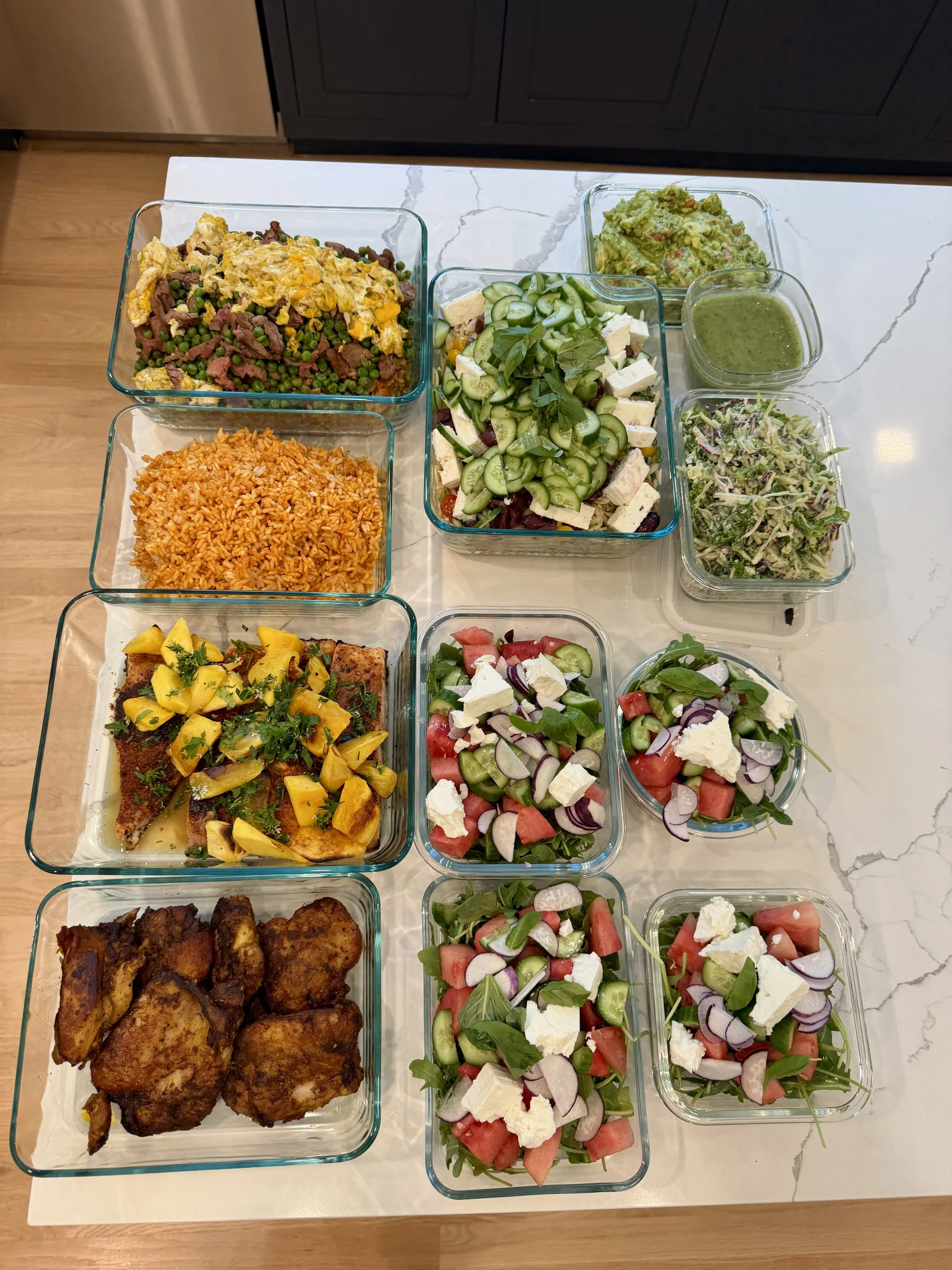 A variety of dishes on a white marble countertop, including fried chicken, rice, salads with cucumbers, tomatoes, and feta cheese, guacamole, and green sauce.