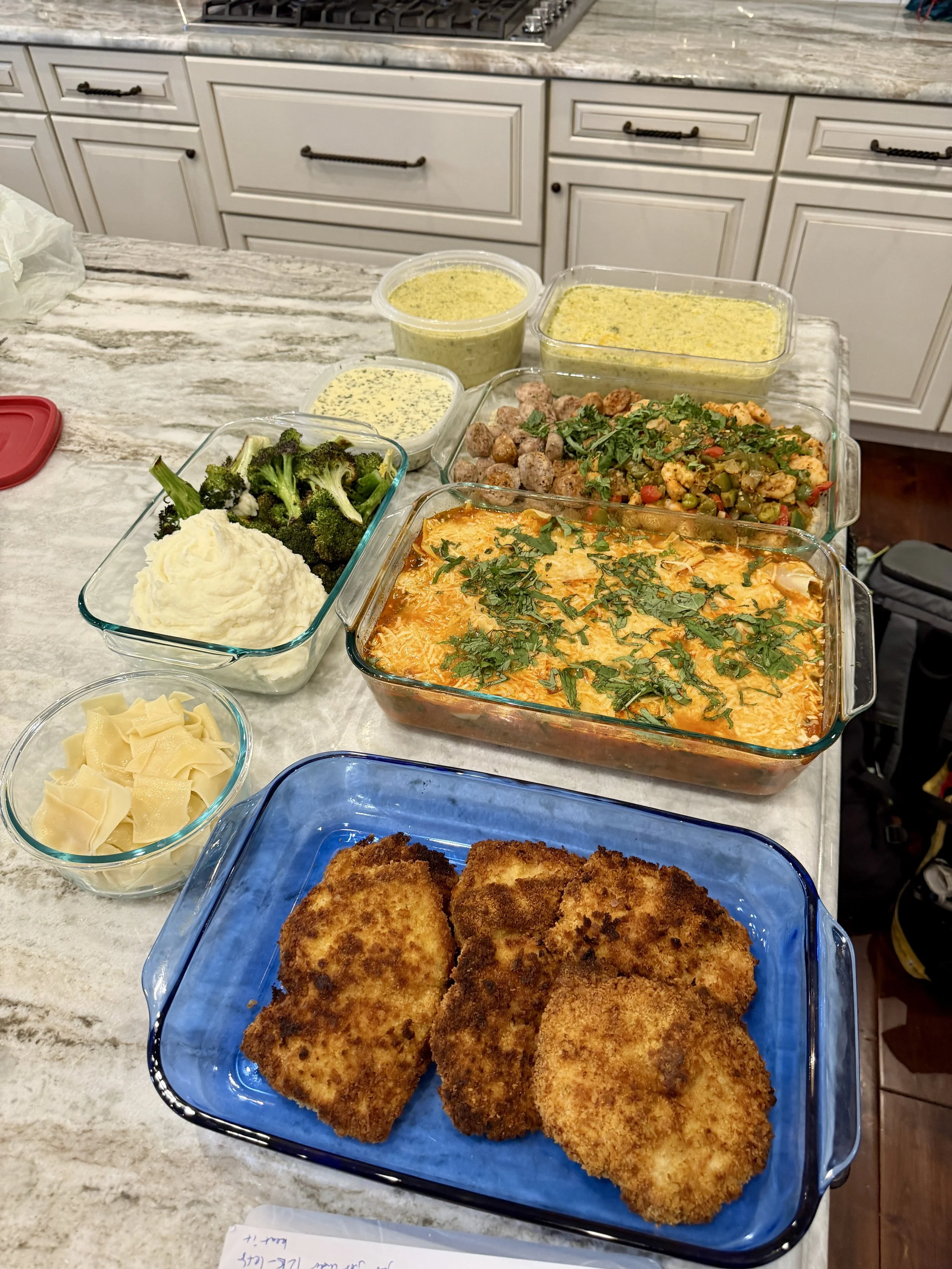 An assortment of homemade dishes on a kitchen counter, including breaded fried items, broccoli, mashed potatoes, shredded cheese, cooked vegetables, meat and vegetable casserole, and containers of green sauce or dip.