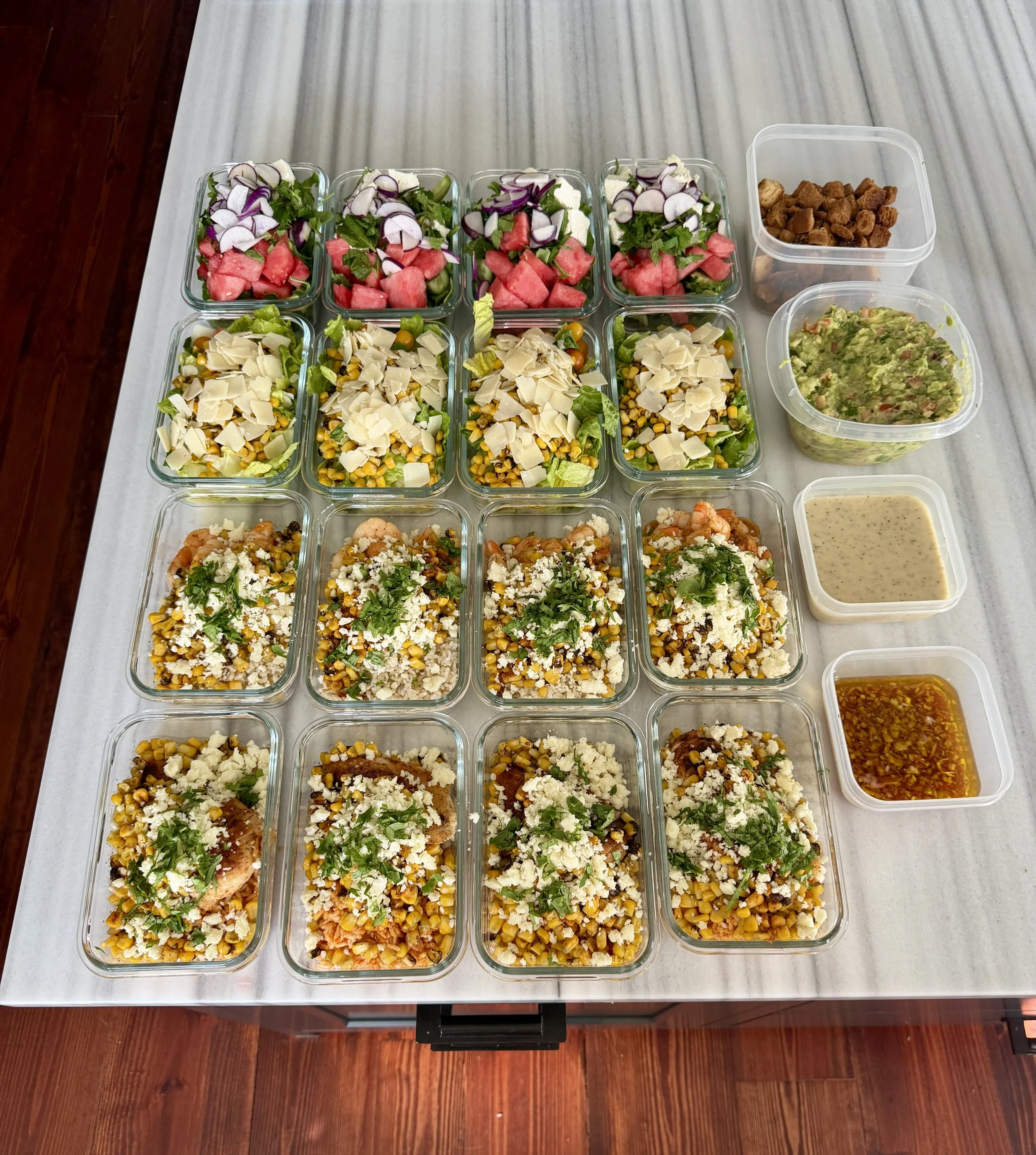 Arrangement of glass food containers on a white table with salads, a container of cooked meat, guacamole, and dressing.