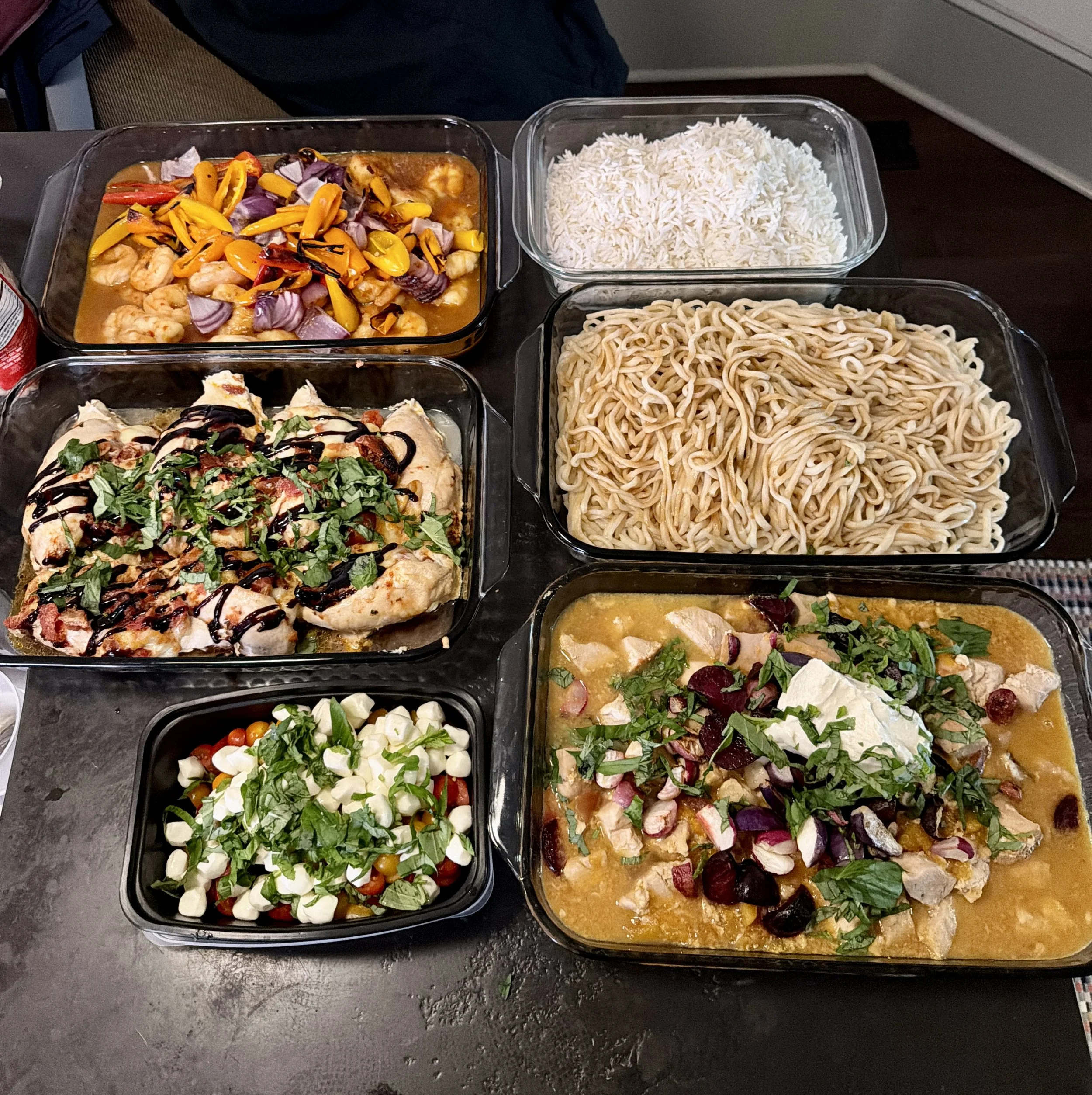 Multiple glass containers filled with various types of food, including pasta, rice, shrimp, pizza, and salads on a dark surface.