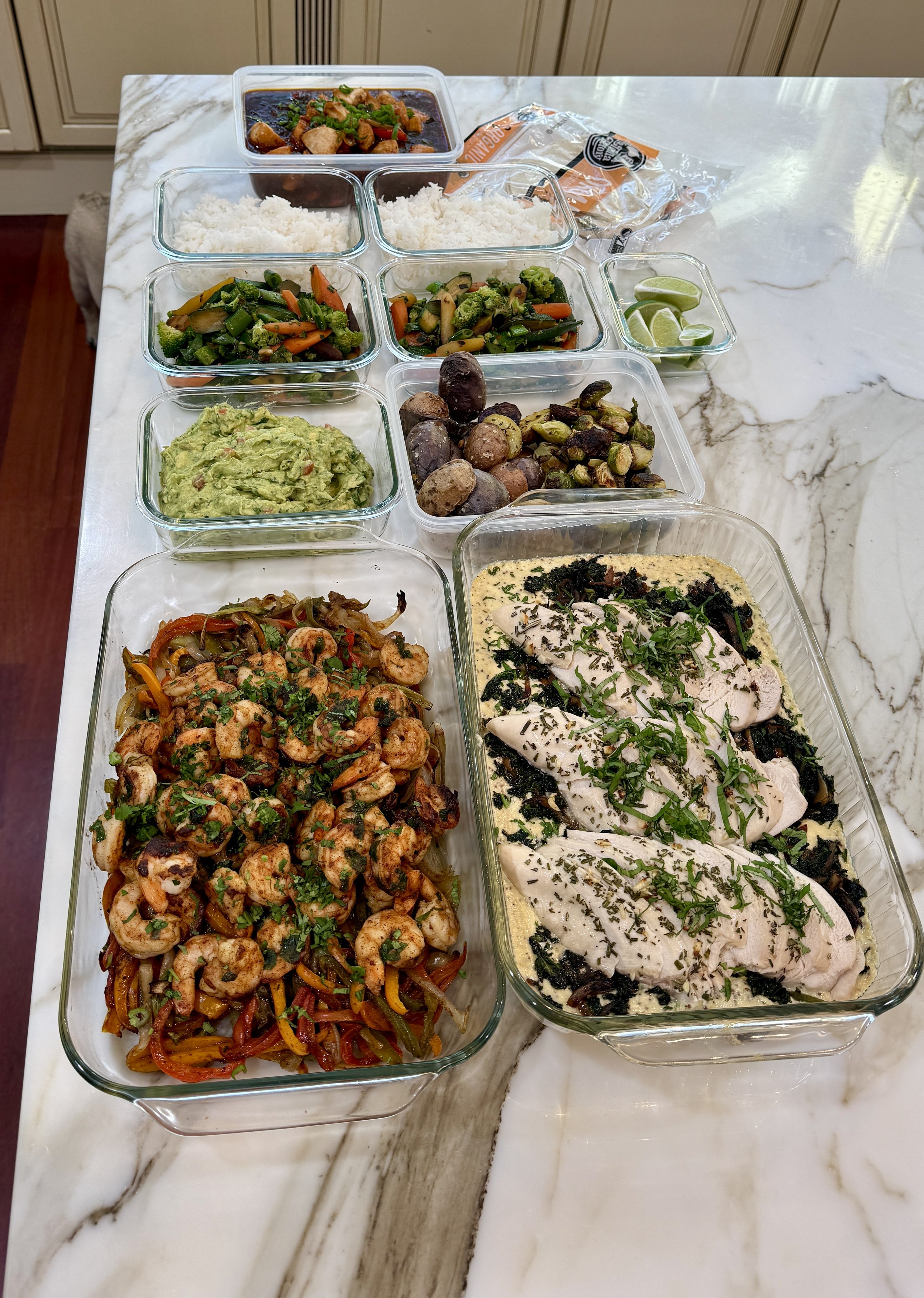 Assorted cooked dishes in glass containers on a white marble countertop, including rice, vegetables, grilled fish, and guacamole, with lime wedges and sliced chili peppers.