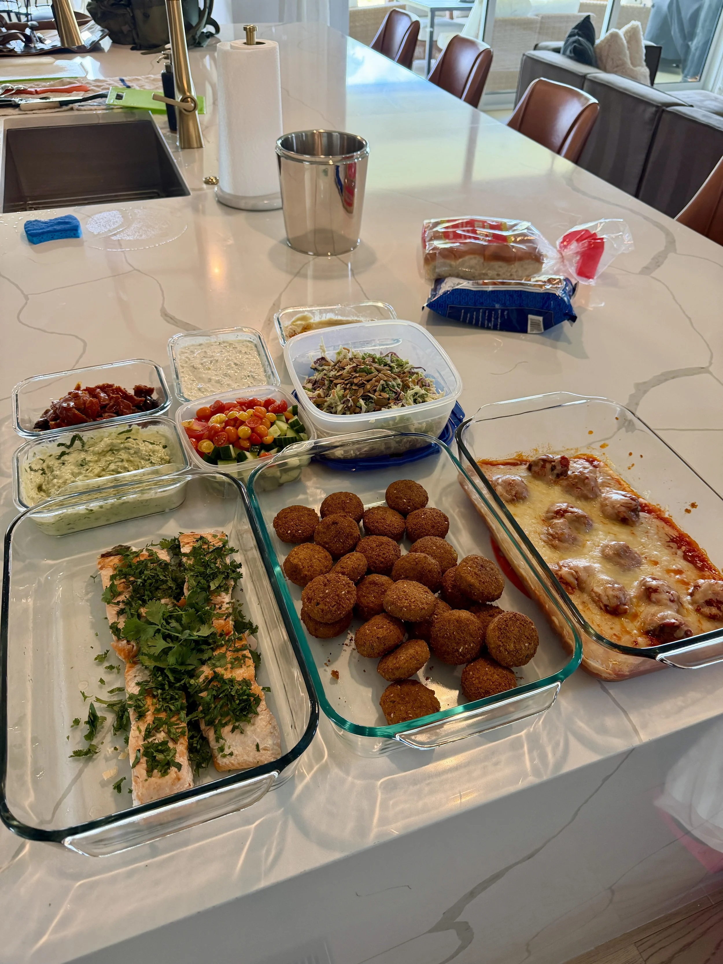 A kitchen countertop holds various dishes including a salmon fillet with herbs, falafel, a tray of pizza, and several containers of dips and salads. There are also a roll of paper towels, a stainless steel cup, and a loaf of bread in plastic wrap.