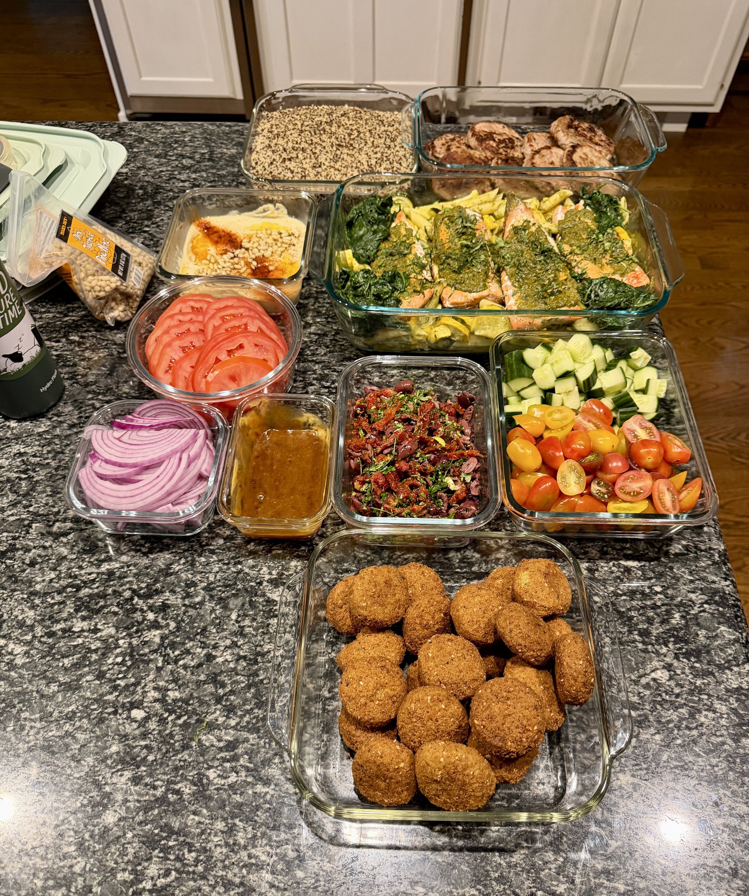 A granite countertop with various glass dishes and containers of food, including sliced tomatoes, red onions, falafel, cooked chicken with green sauce, ground sesame seeds, and a creamy dip with spices.
