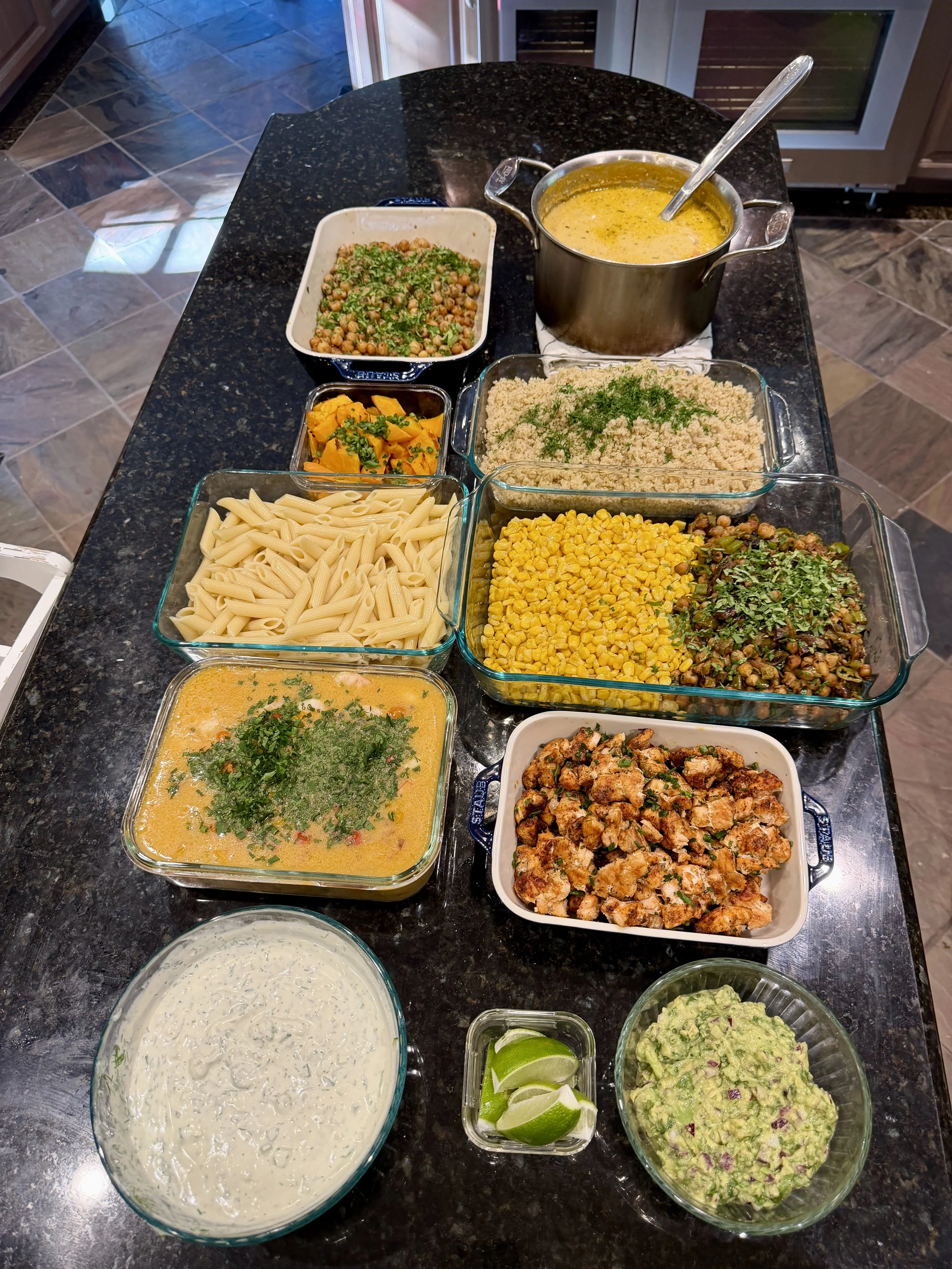 A variety of prepared food dishes on a black granite kitchen countertop, including pasta, various vegetable dishes, and dips.