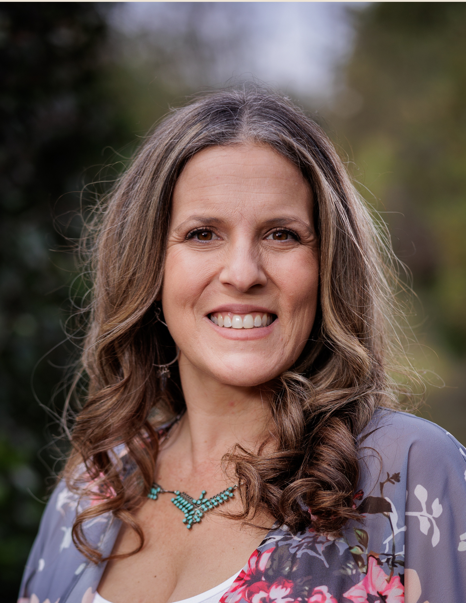 Smiling person with long, wavy hair wearing a floral outfit and turquoise necklace, outdoors