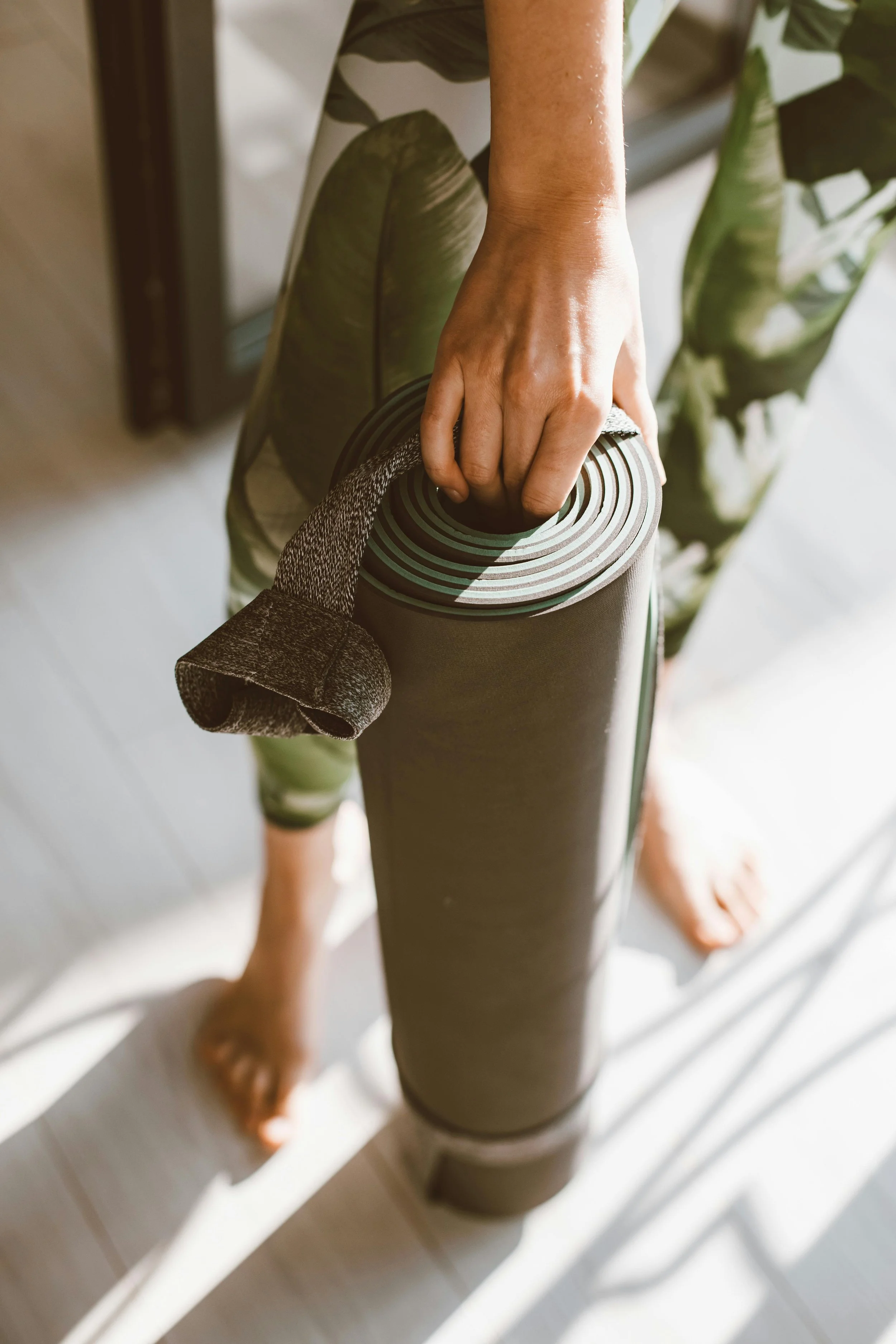 Person holding rolled yoga mat with a strap, wearing green leaf-patterned leggings.